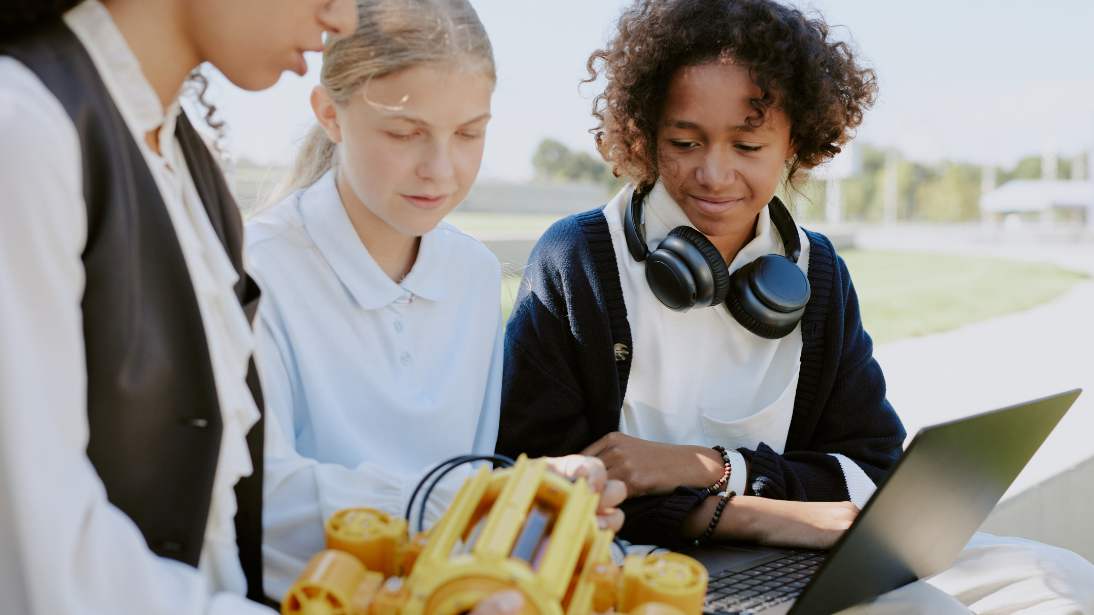 Three students looking at a laptop outdoors on a sunny day, with yellow plastic building blocks in the foreground.