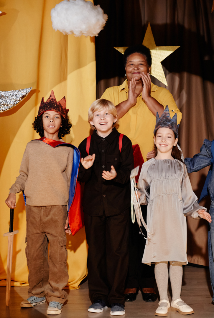 Children dressed in costume on stage with a woman, stars and clouds hanging overhead, during a celebration or performance.