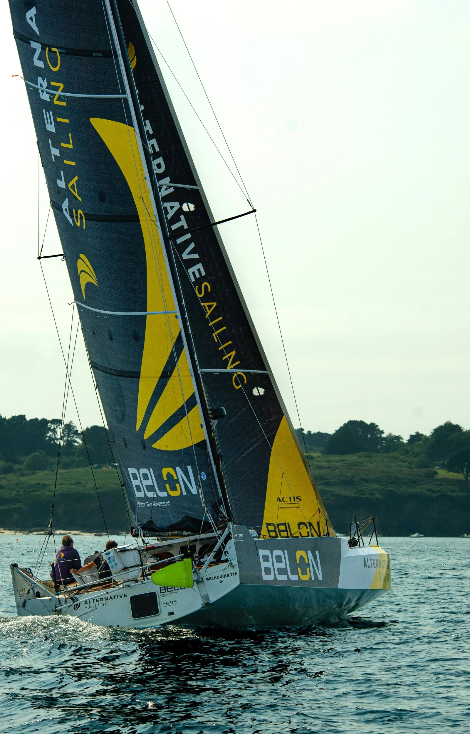 Voilier de course à voile avec voile noire et jaune affichant 'BELON' et 'ALTERNATIVE SAILING', naviguant sur l'eau avec un paysage de collines en arrière-plan.