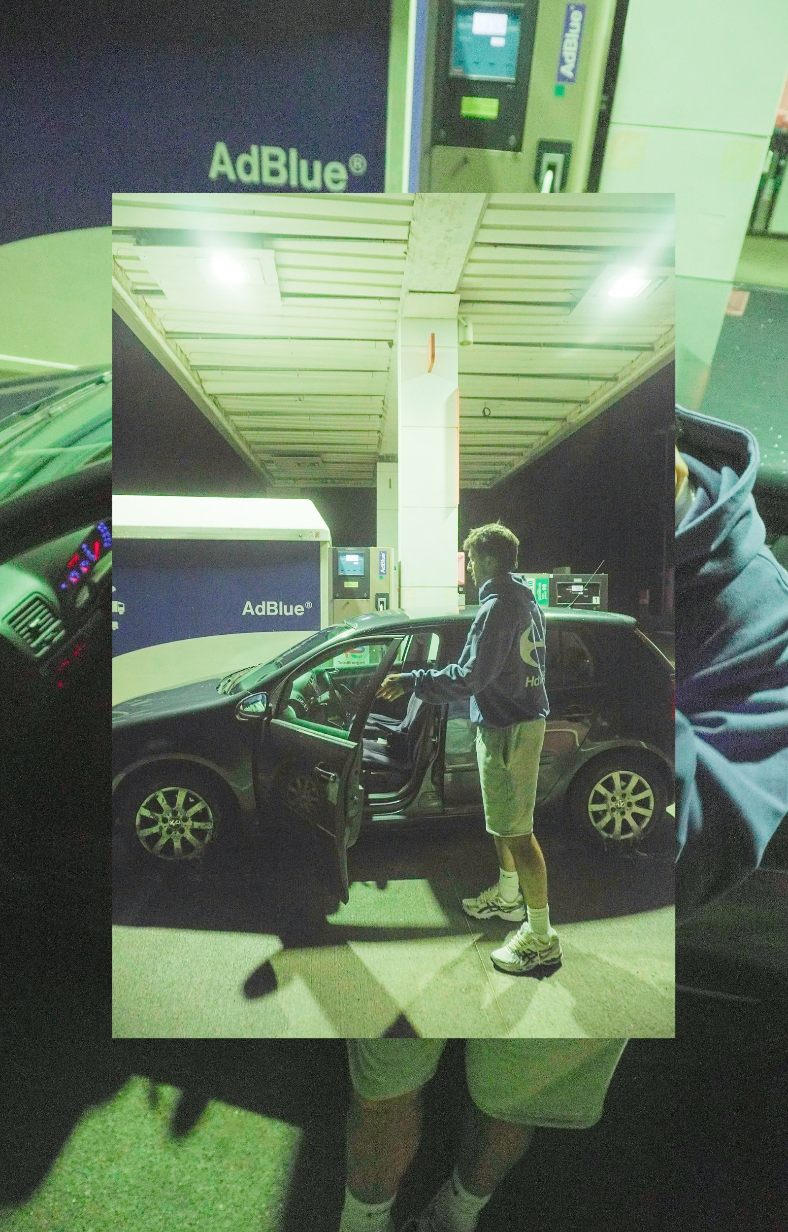 Un jeune homme qui se tient à côté d'une voiture, portant un sweat à capuche, un short et des baskets, dans une station-service la nuit. On voit des écrans de distributeurs automatiques derrière lui.