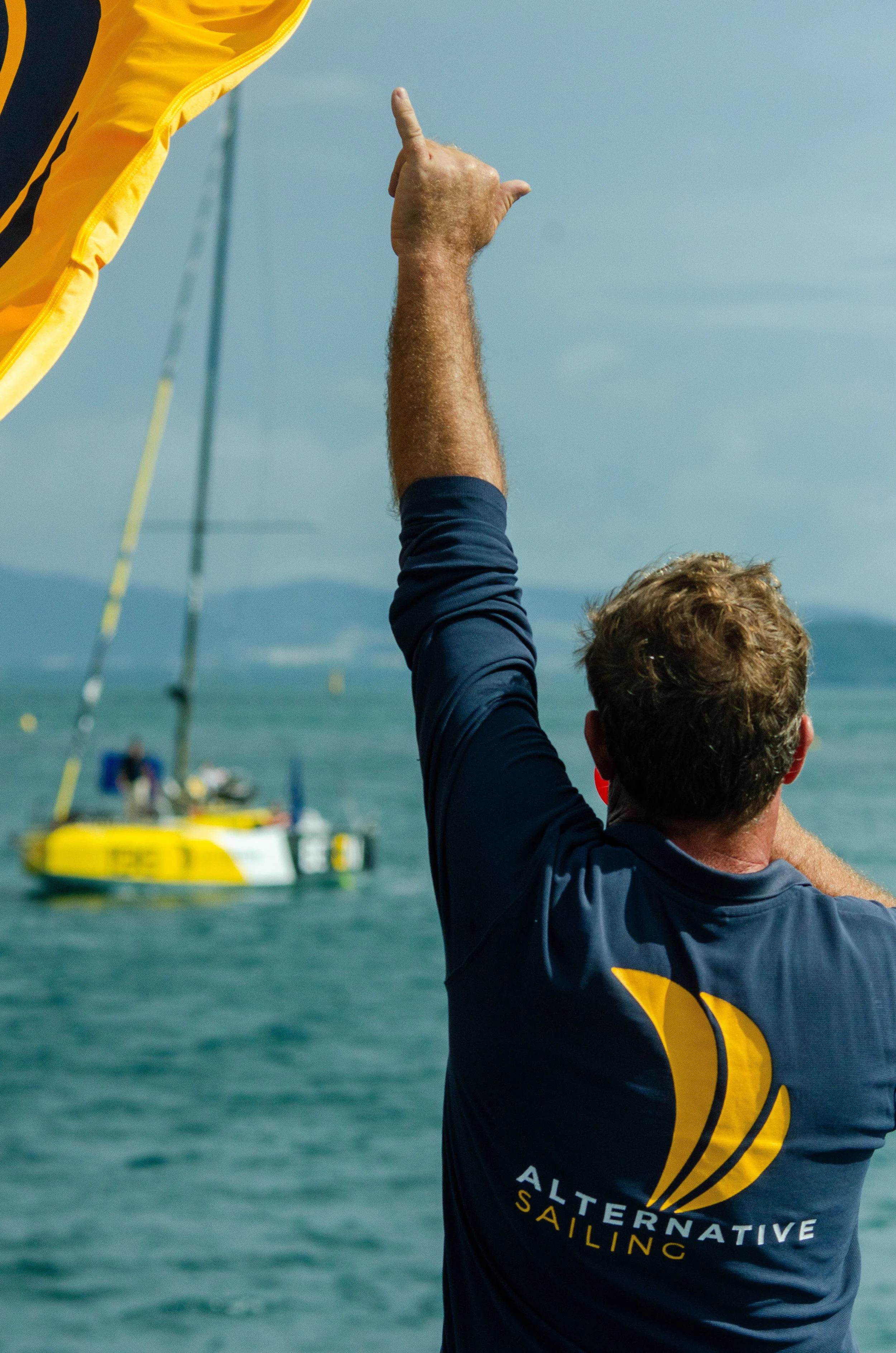 Un homme avec des cheveux bouclés, vêtu d'un maillot de sport bleu marine avec un logo jaune 'ALTERNATIVE SAILING' au dos, qui fait un geste de victoire avec le bras levé, sur fond de mer avec voilier visible en arrière-plan.