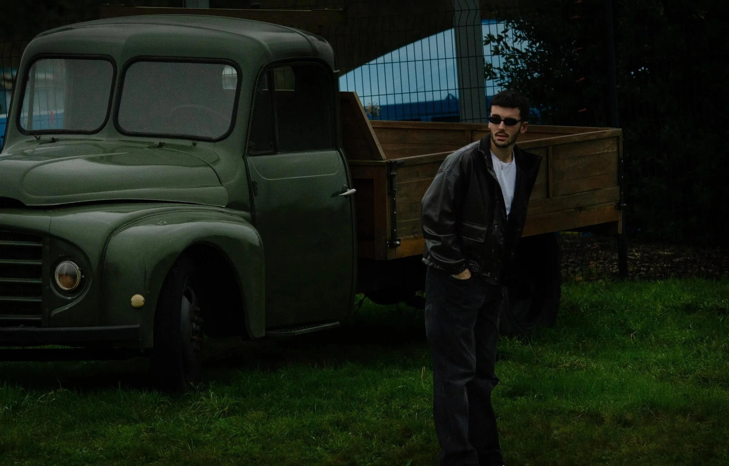 Jeune homme portant des lunettes de soleil, vêtu d'une veste noire et d'un t-shirt blanc, debout à côté d'un camion en bois vert foncé, dans un jardin ou un espace extérieur clôturé.