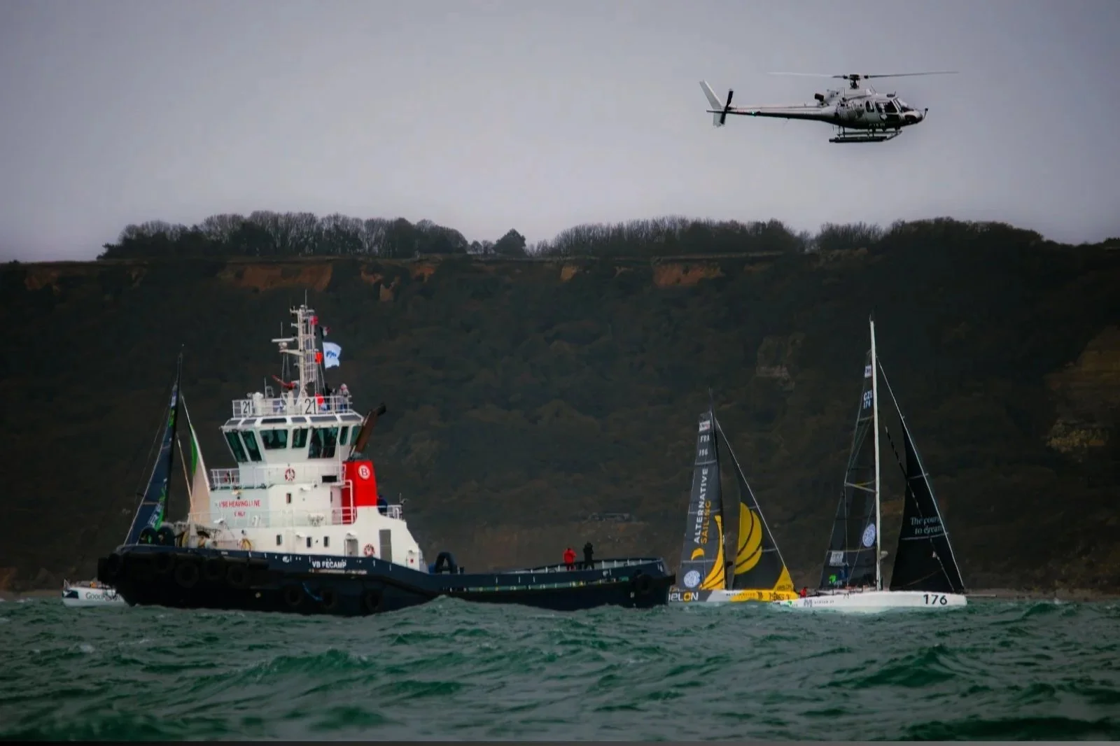 Un bateau de remorquage tirant un voilier de course dans des eaux agitées avec une falaise en arrière-plan et un hélicoptère volant dans le ciel gris.