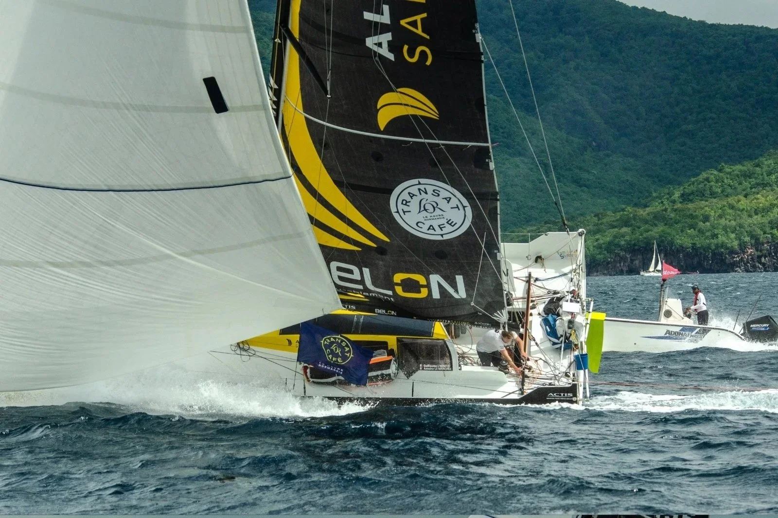 Voilier en compétition sur la mer avec voile noir et jaune, d'autres bateaux en arrière-plan, montagnes verdoyantes au loin.