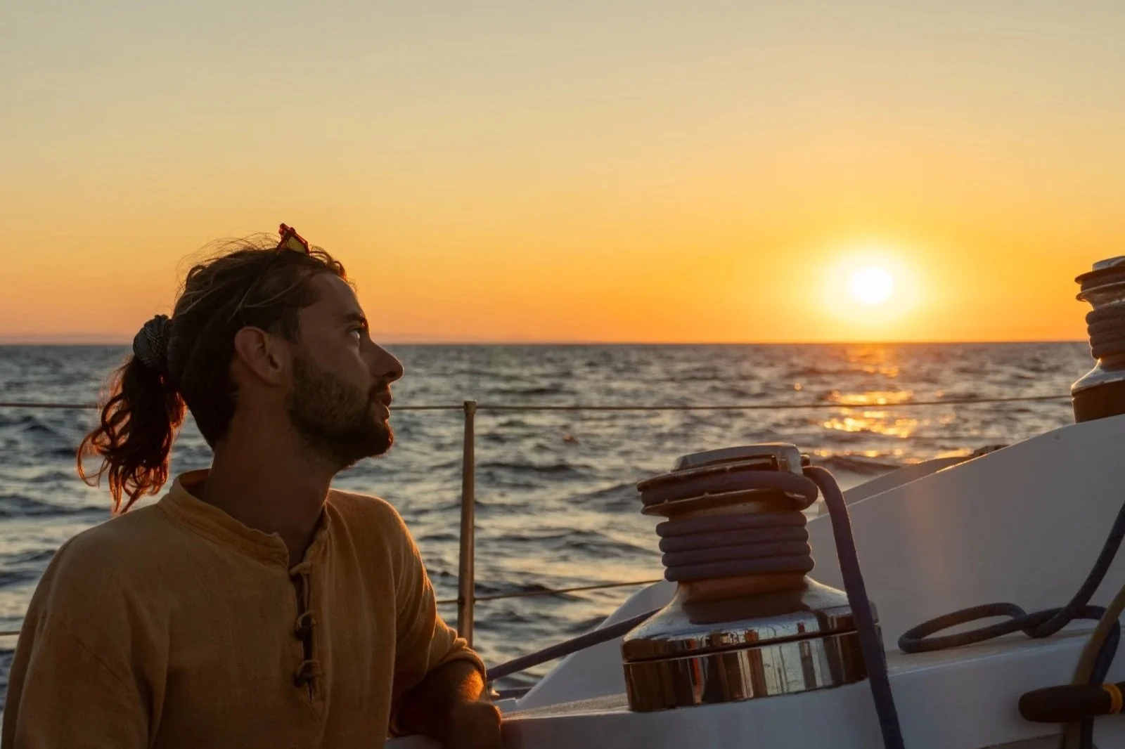 Homme avec cheveux longs attachés, regardant vers le coucher du soleil depuis un bateau en mer, avec des cordages et des équipements de navigation visibles.