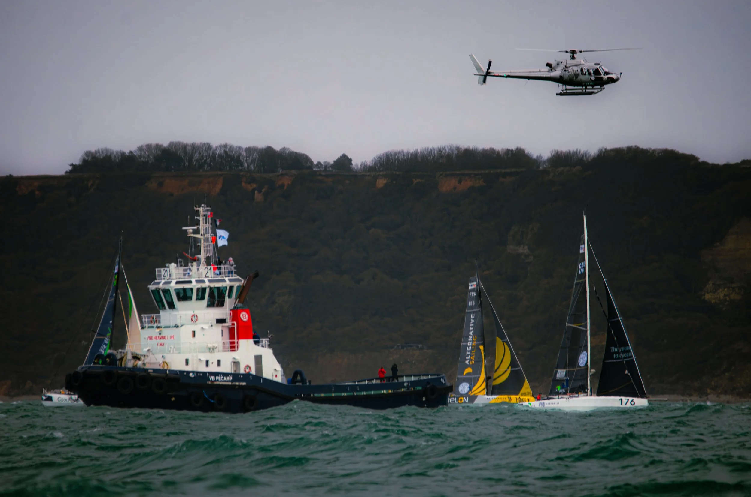 Un bateau de remorquage en mer avec deux voiliers à côté et un hélicoptère volant au-dessus, avec des collines en arrière-plan.