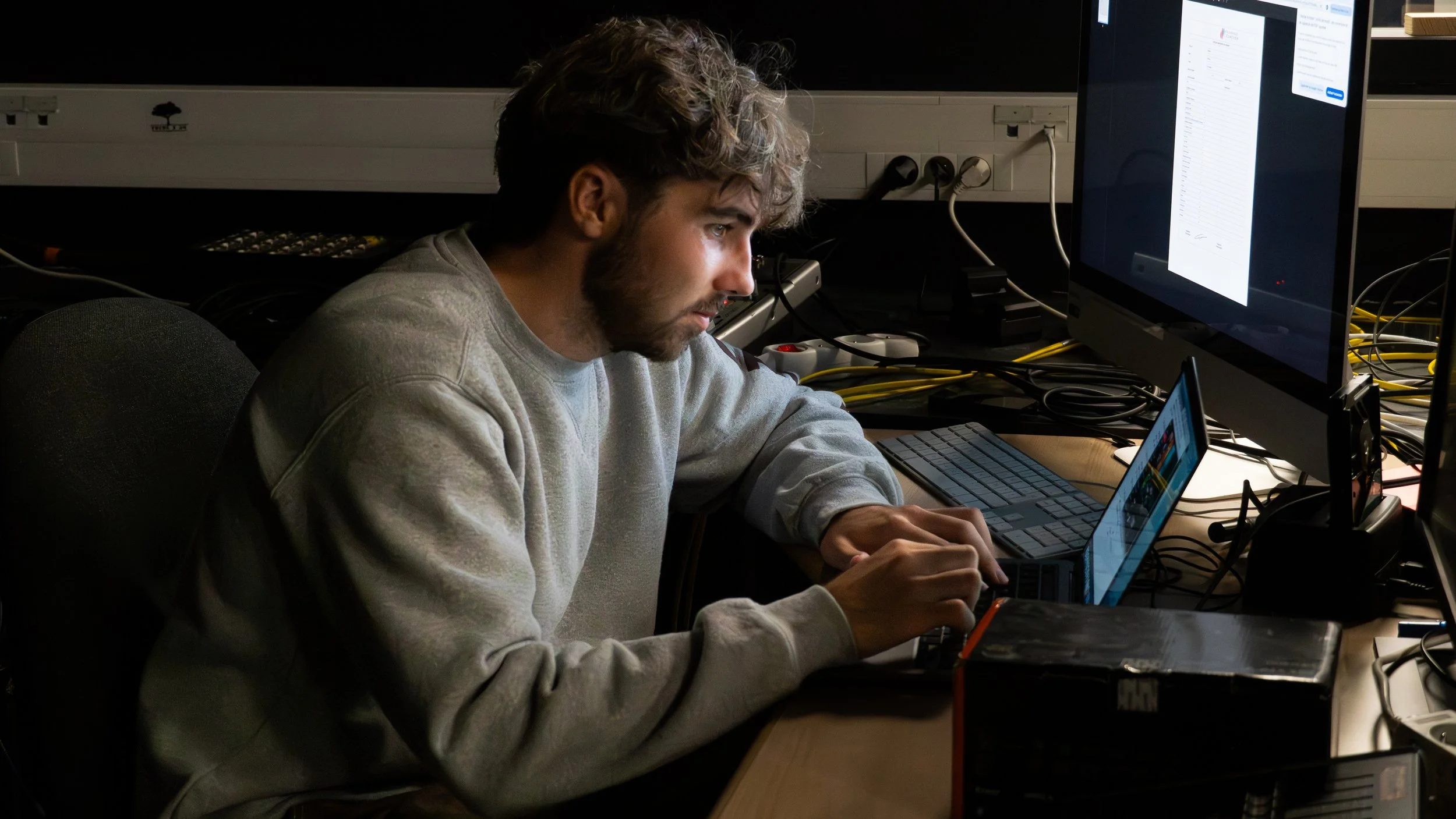 Un jeune homme travaillant sur un ordinateur portable dans un bureau sombre, avec plusieurs écrans et câbles autour.
