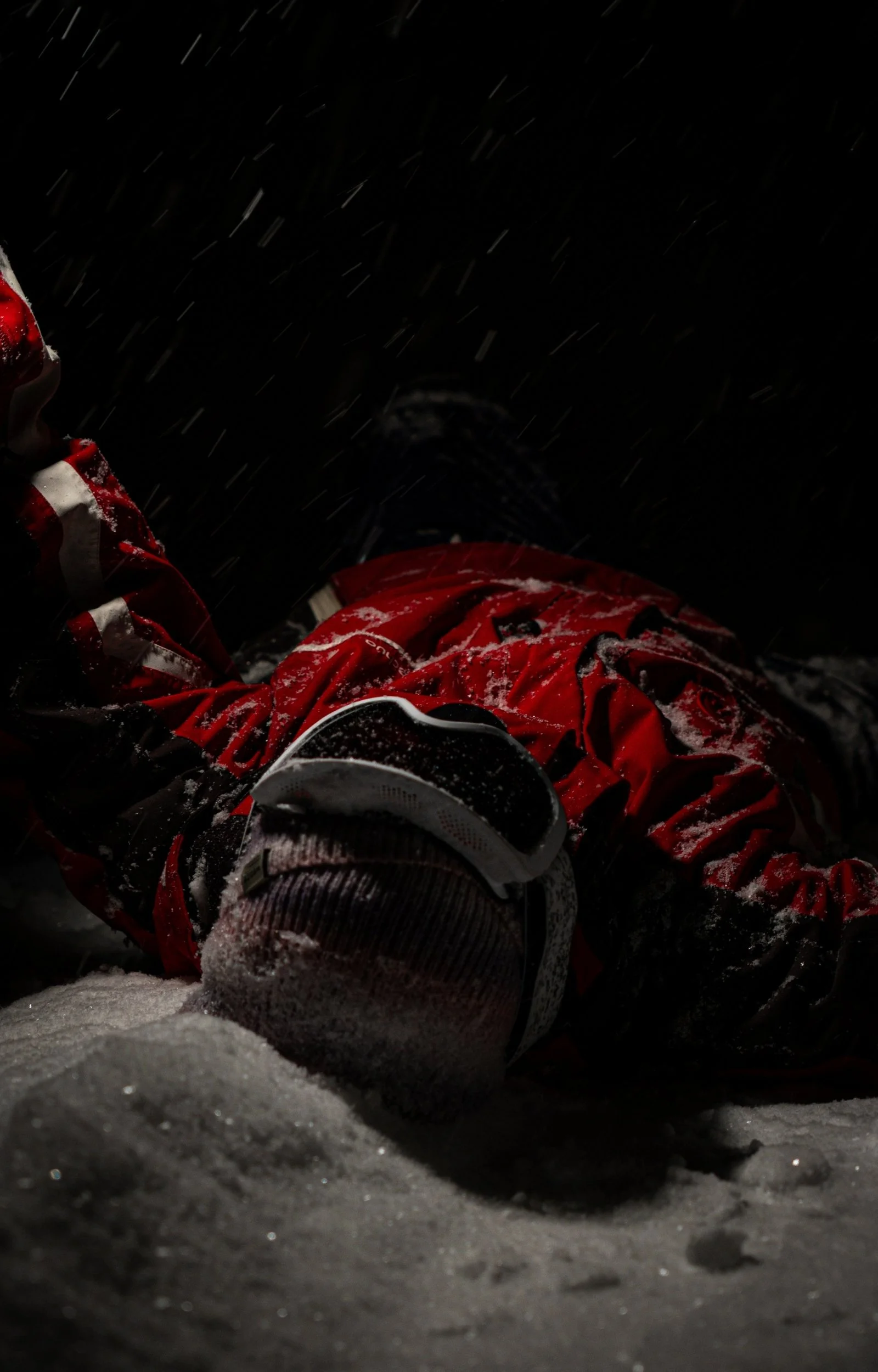 Personne allongée dans la neige la nuit, portant des vêtements de sport rouges, avec de la neige qui tombe.