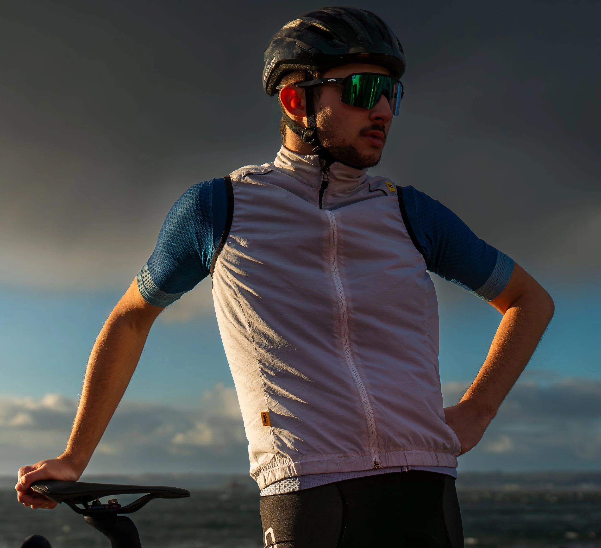 Un homme portant un casque de vélo, des lunettes de soleil teintées, une veste blanche et un t-shirt bleu, se tient avec une main sur le guidon d'une bicyclette, sous un ciel orageux ou nuageux.