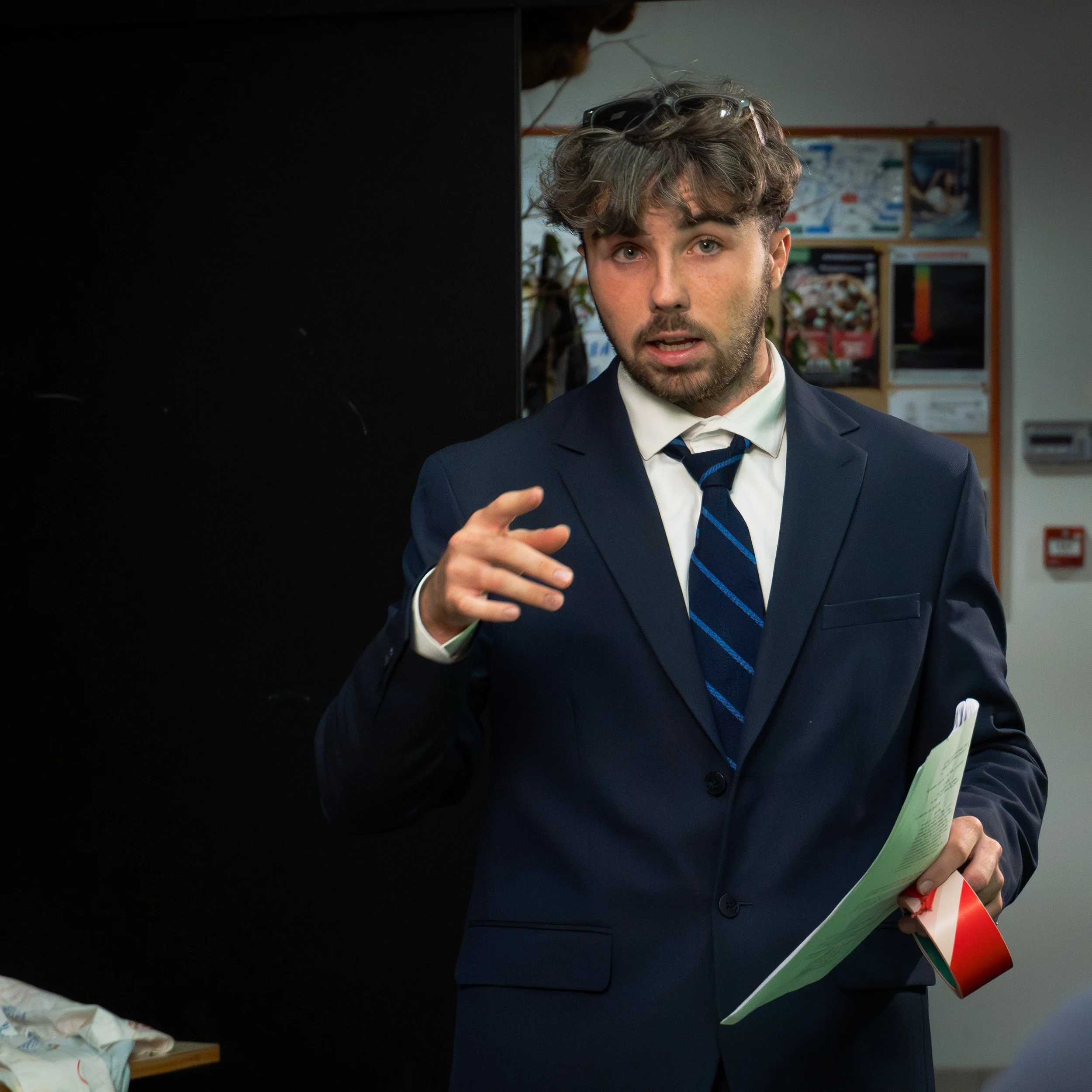 Un homme en costume bleu, avec une chemise blanche et une cravate, tient des documents dans une main et parle ou explique quelque chose avec une expression sérieuse. Il porte des lunettes sur la tête.