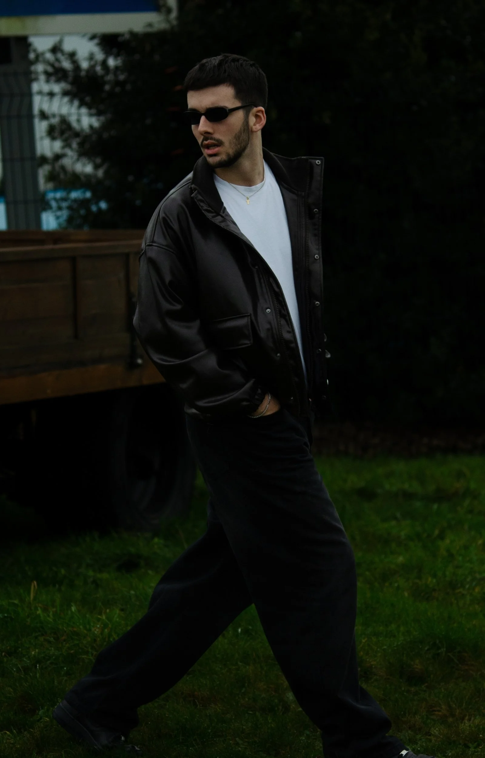 Un jeune homme portant des lunettes de soleil noires, un t-shirt blanc, une veste noire en cuir et un pantalon noir marche dans un jardin ou un espace extérieur avec des arbres et de l'herbe verte.