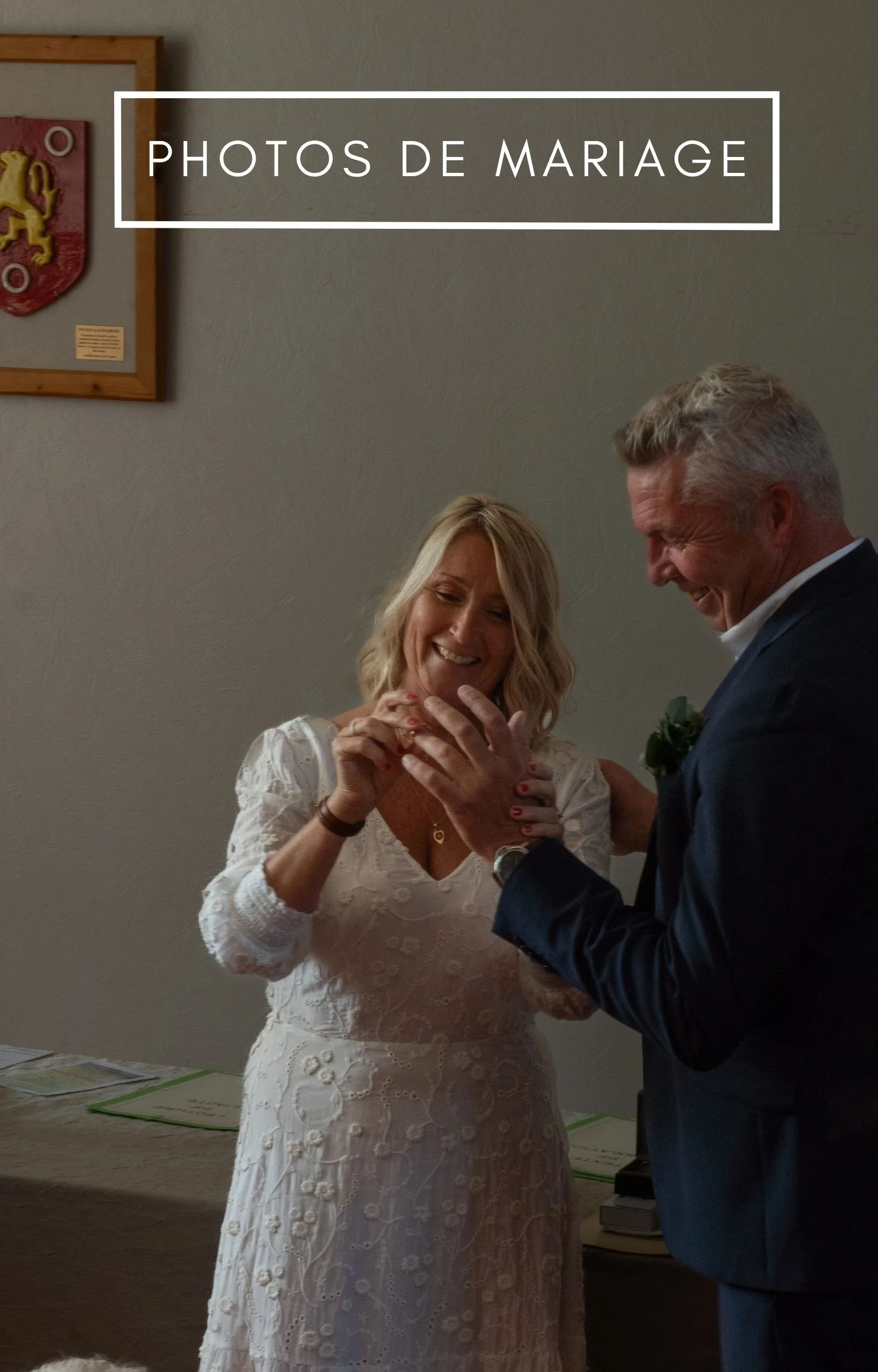 Une femme en robe blanche se marie, échange des alliances avec un homme en costume noir, tous deux souriants, dans une salle avec mur beige et un tableau à l'arrière.