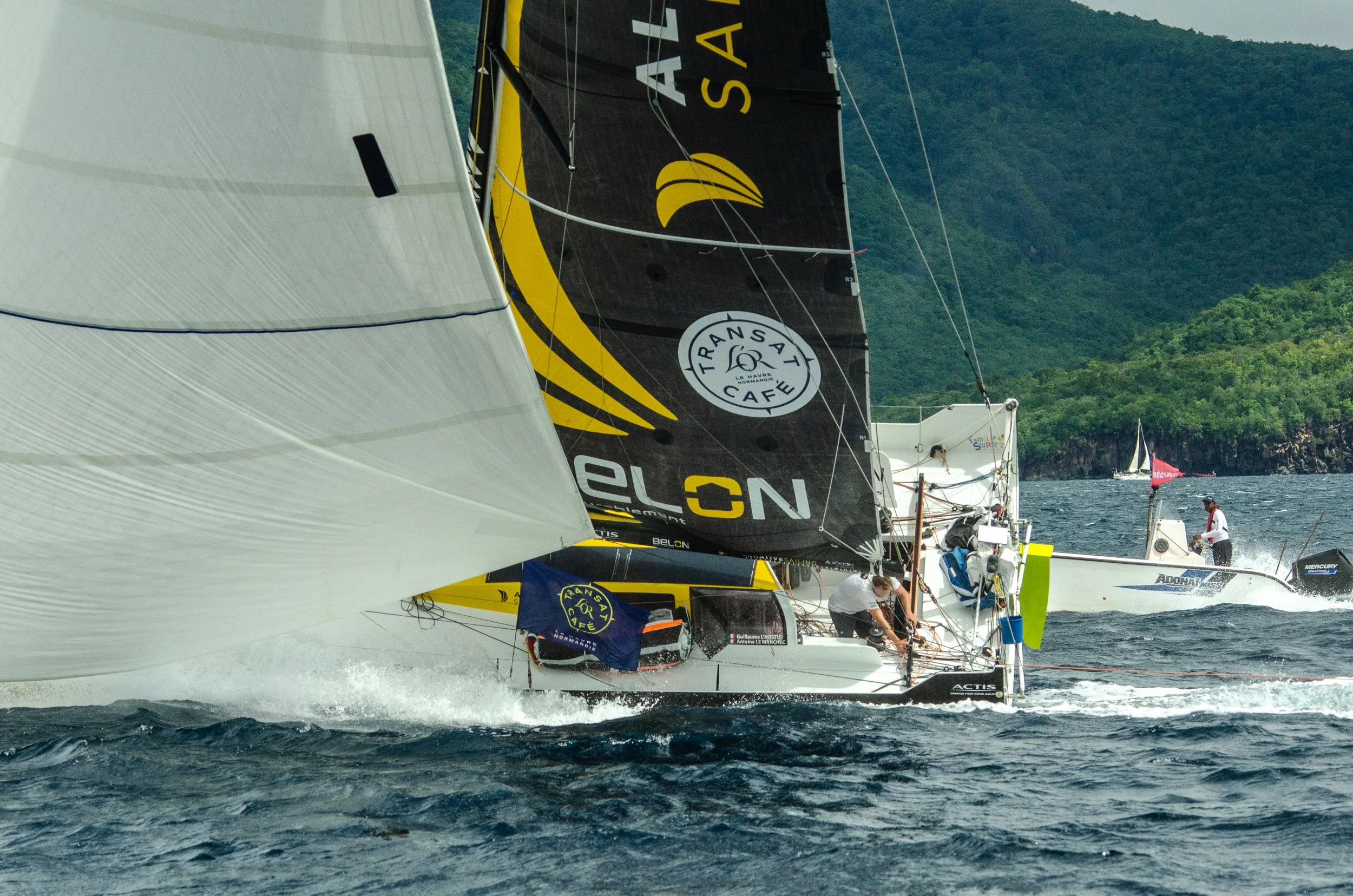 Voiliers de course naviguant en mer avec un paysage montagneux en arrière-plan.