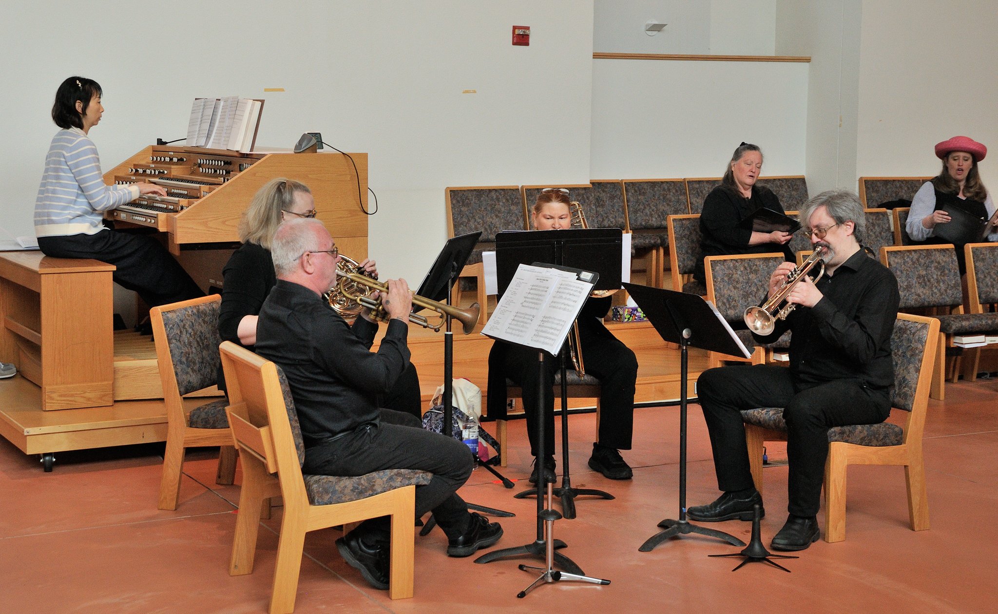 Six musicians performing in a room, including a woman on the organ, a man and woman playing trumpets, and three women singing, two holding black folders, one wearing a pink hat.