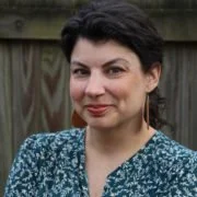 Portrait of a woman with short dark hair and a floral blouse, standing outdoors in front of a wooden fence.