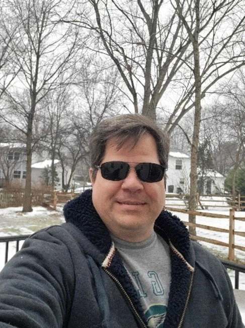Man wearing sunglasses and a hoodie takes a selfie outdoors in a winter neighborhood with snow-covered ground and leafless trees.
