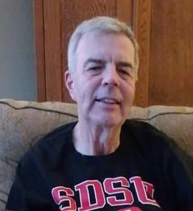 A man with gray hair sitting on a beige couch, wearing a black T-shirt with red and white text.