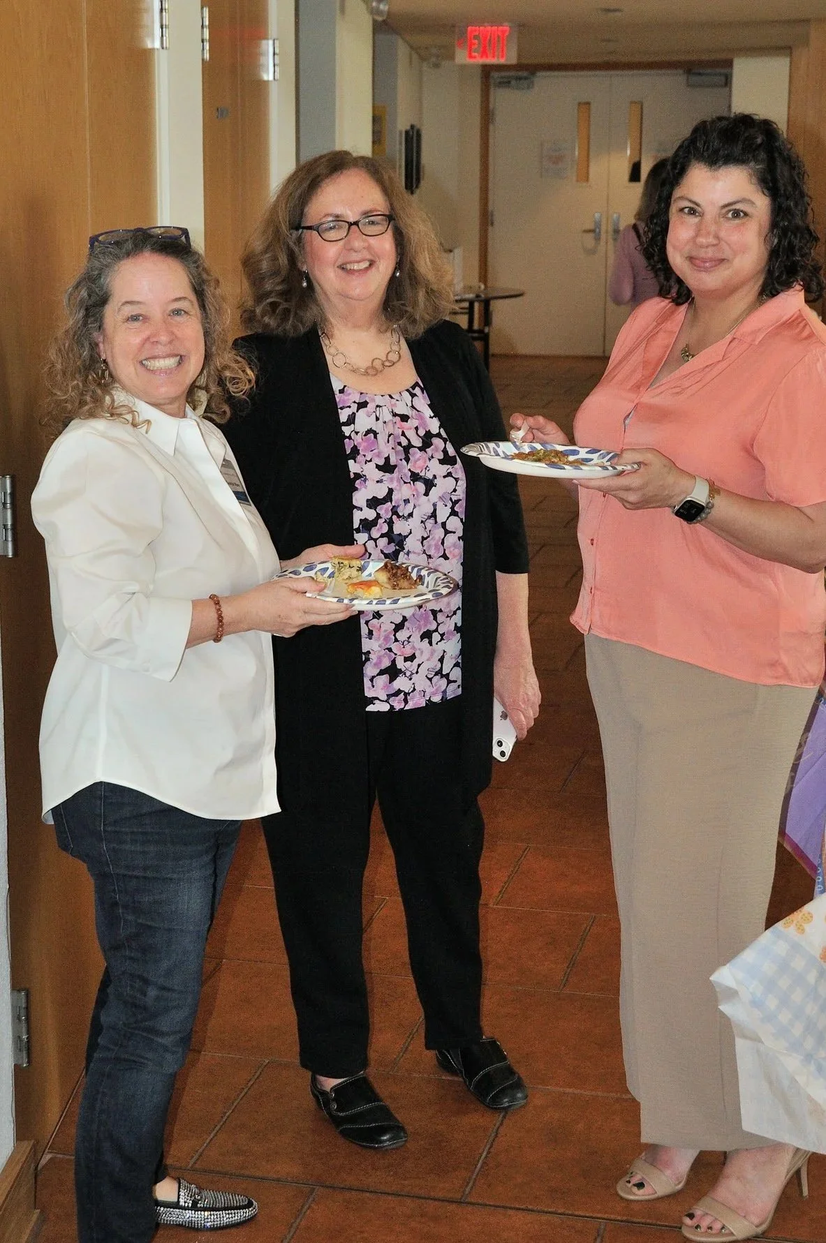 Three women standing indoors, smiling, holding plates of food at a gathering or event.