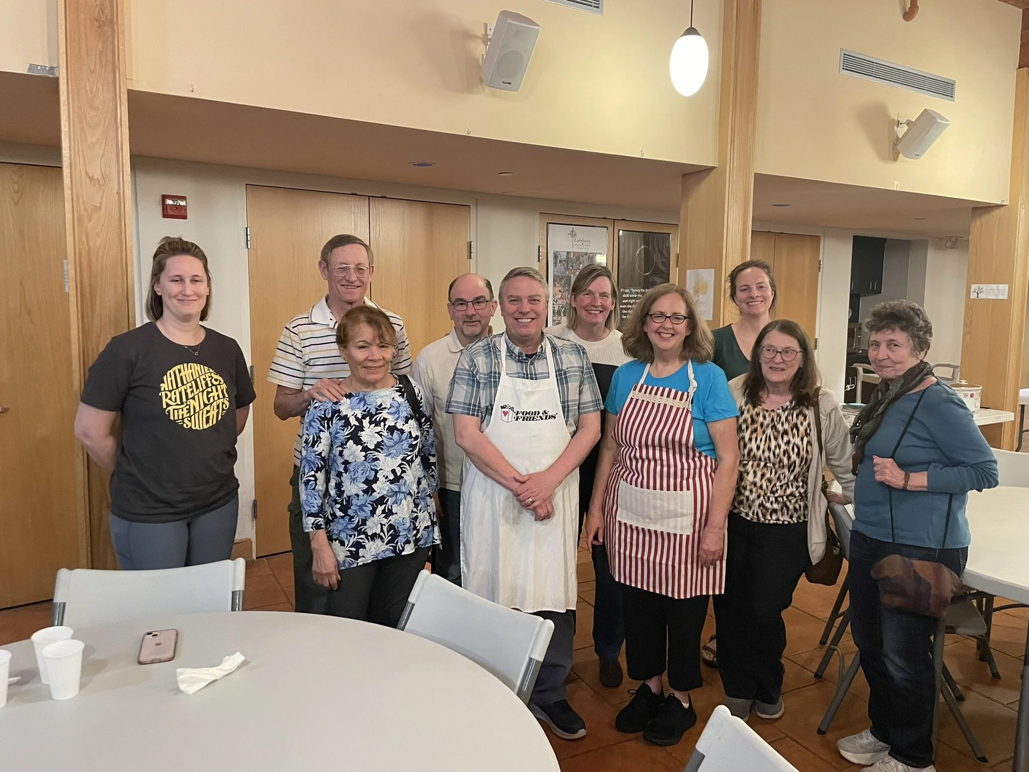 Group of people gathered in a room, some wearing aprons, suggesting a cooking class or community event.