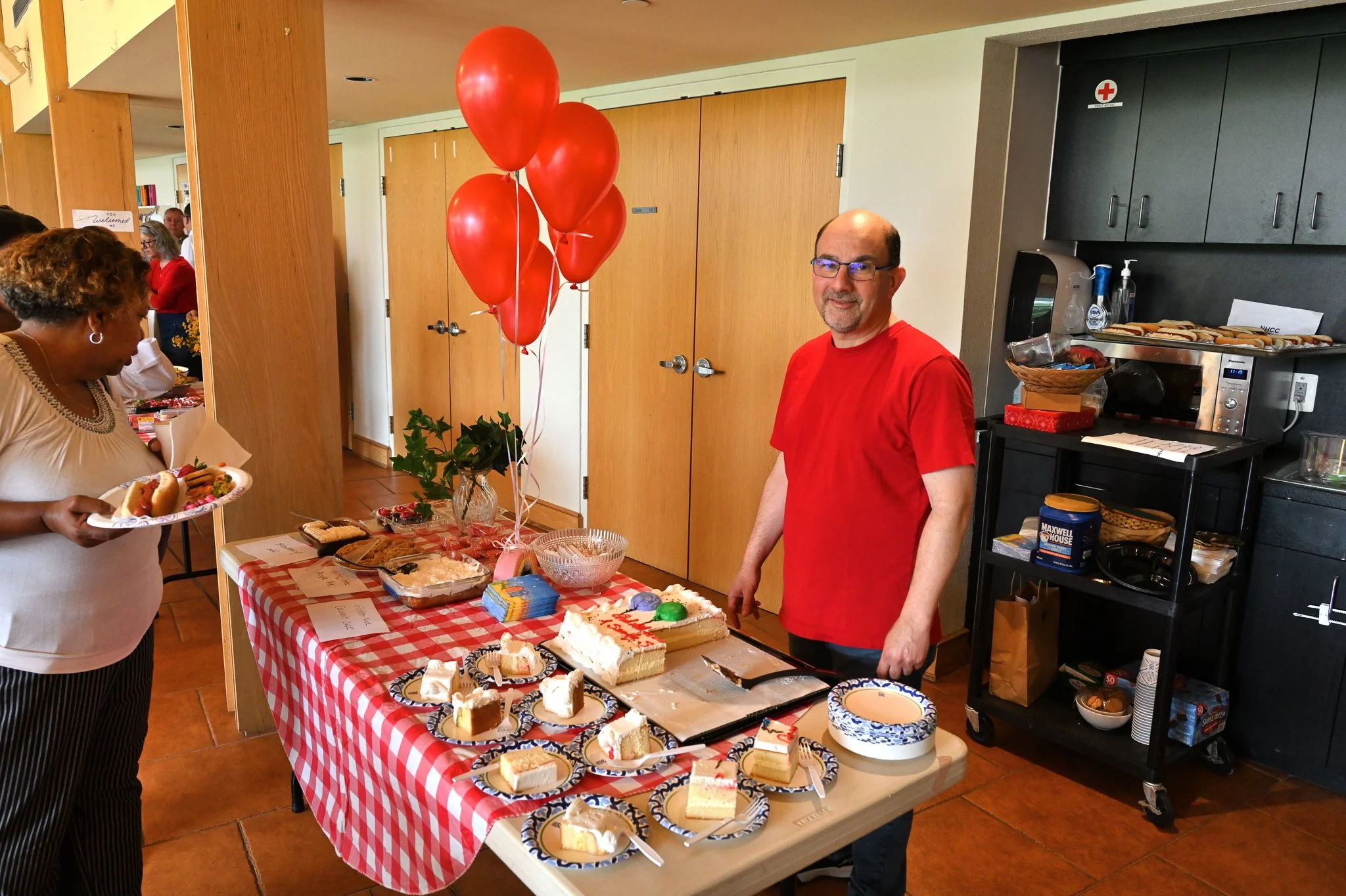 People at a celebration, with a table of desserts including cake slices, cookies, and layered cakes, decorated with red balloons, in a room with wooden doors and a kitchen area.