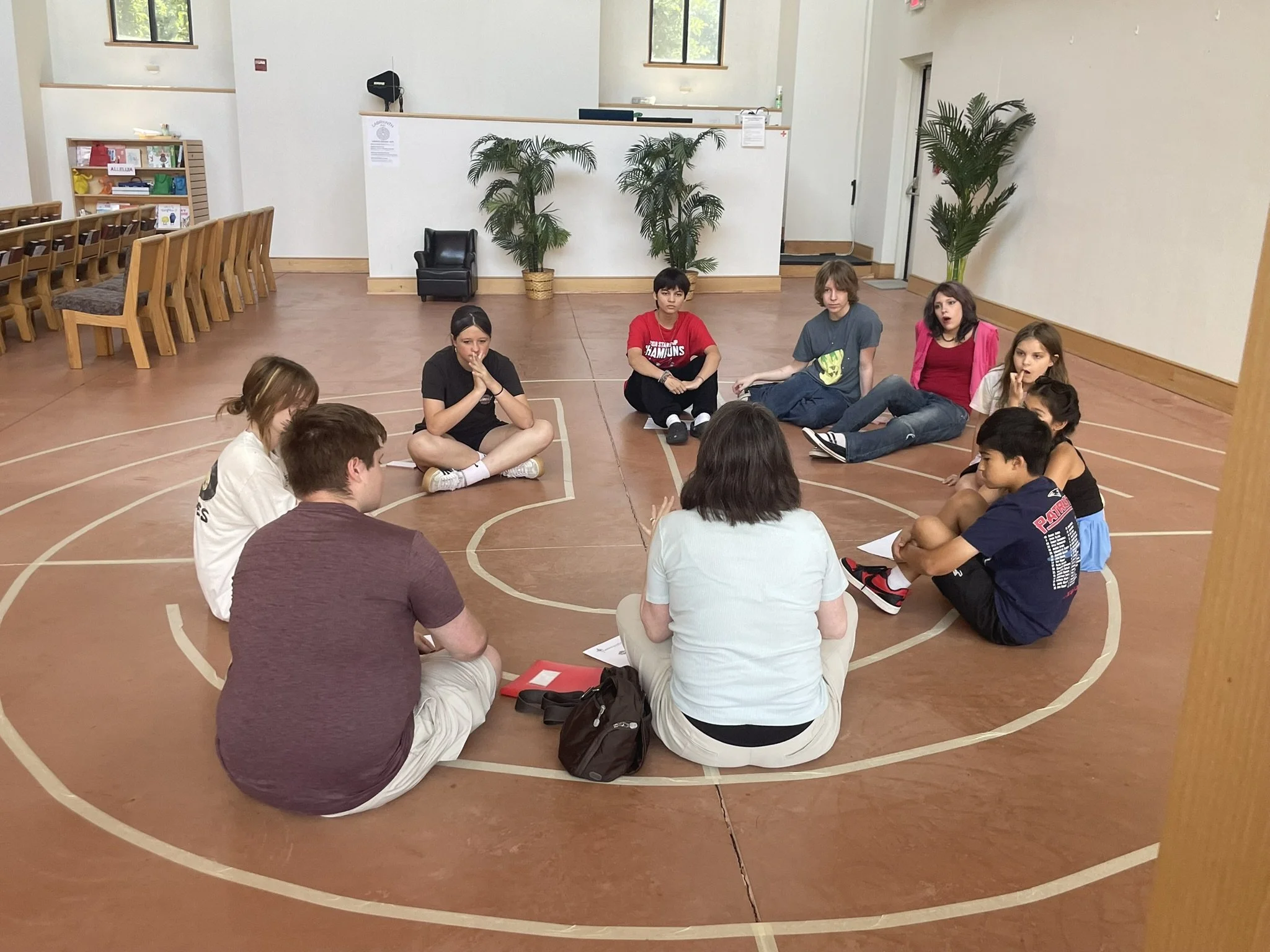 A group of children sitting cross-legged in a circle on a wooden gymnasium floor, with some holding papers. The background includes potted plants, windows, and a small black chair.