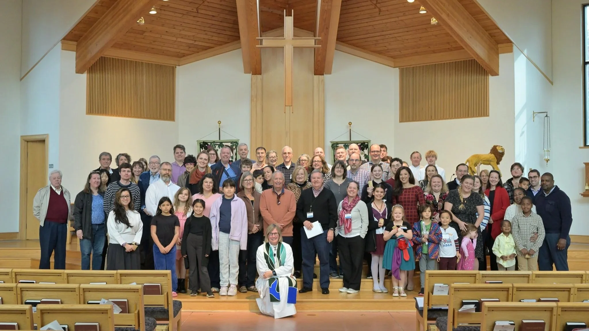 A large group of people, including children and adults, gathered on the stage of a church or community center with wooden architecture, posing for a group photo.