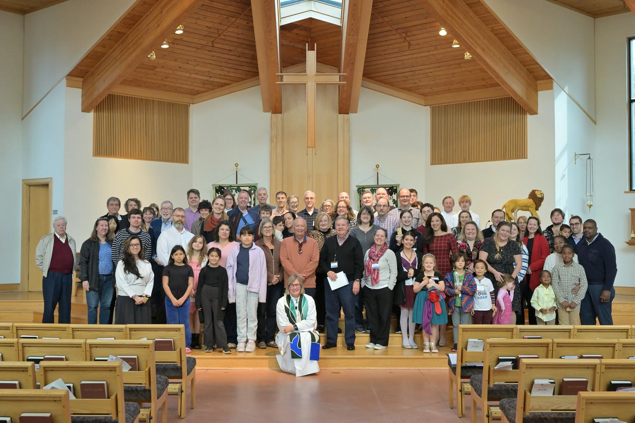 A large group of people, including children and adults, gathered on the stage of a church or auditorium for a group photo. The setting has a wooden ceiling with beams and a cross at the center.