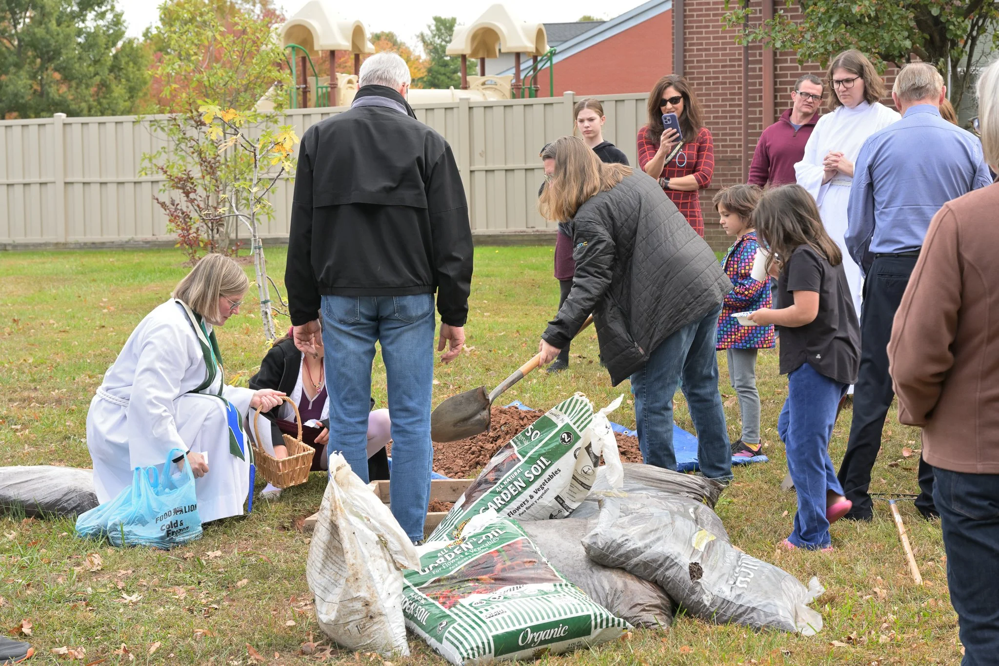 People planting a young tree in a backyard, surrounded by gardening supplies like soil bags, with a fence, trees, and a playset in the background.