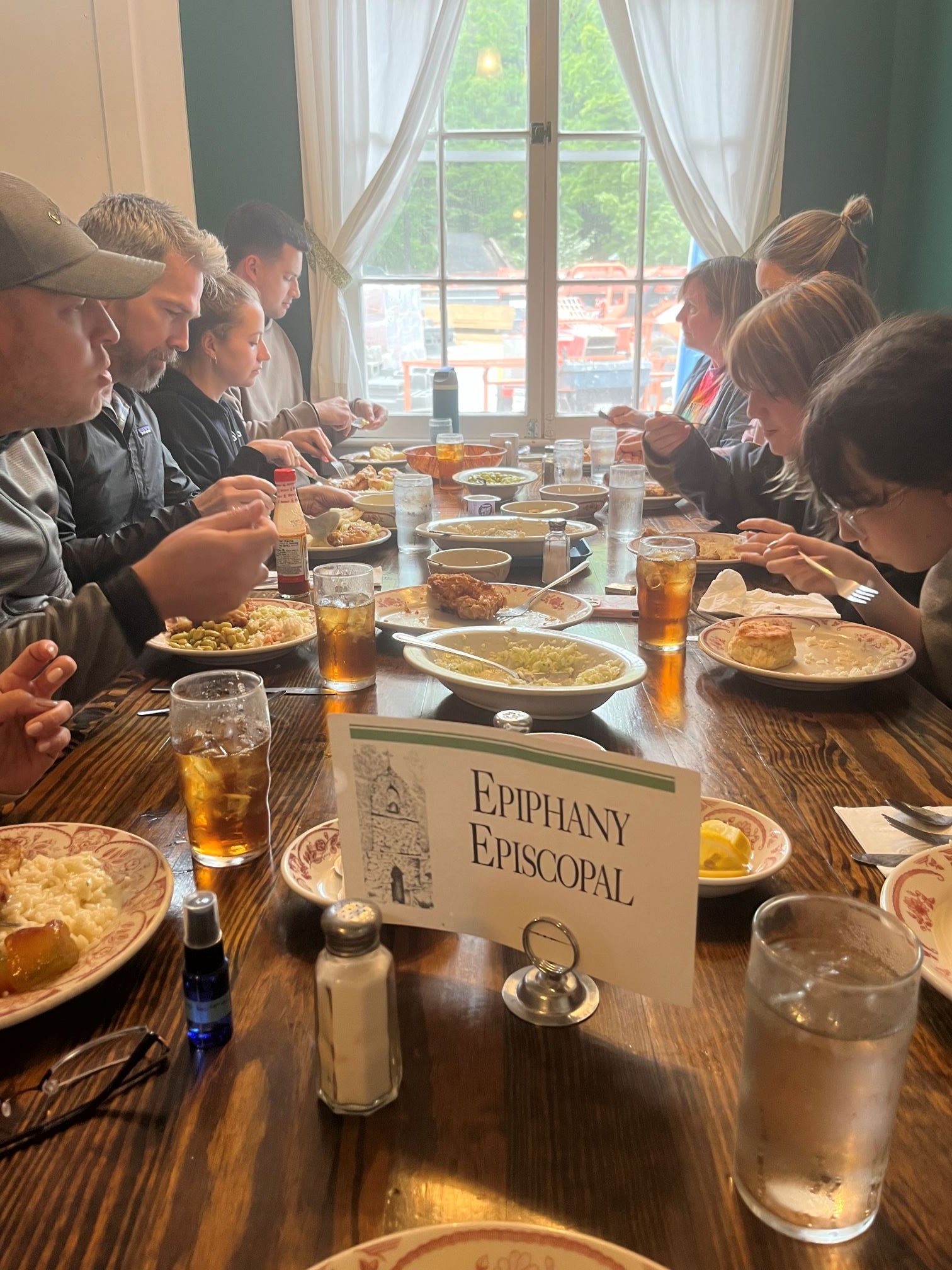 People seated at a wooden table having a meal in a cozy restaurant with a large window overlooking a green outdoor area. Plates of food, glasses of iced tea, and condiments are on the table. A sign with the words 'Epiphany Episcopal' is in the foregr