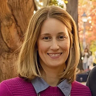 A woman with shoulder-length blonde hair smiling outdoors, wearing a purple sweater and a denim collar shirt, with a tree and blurred autumn scenery in the background.