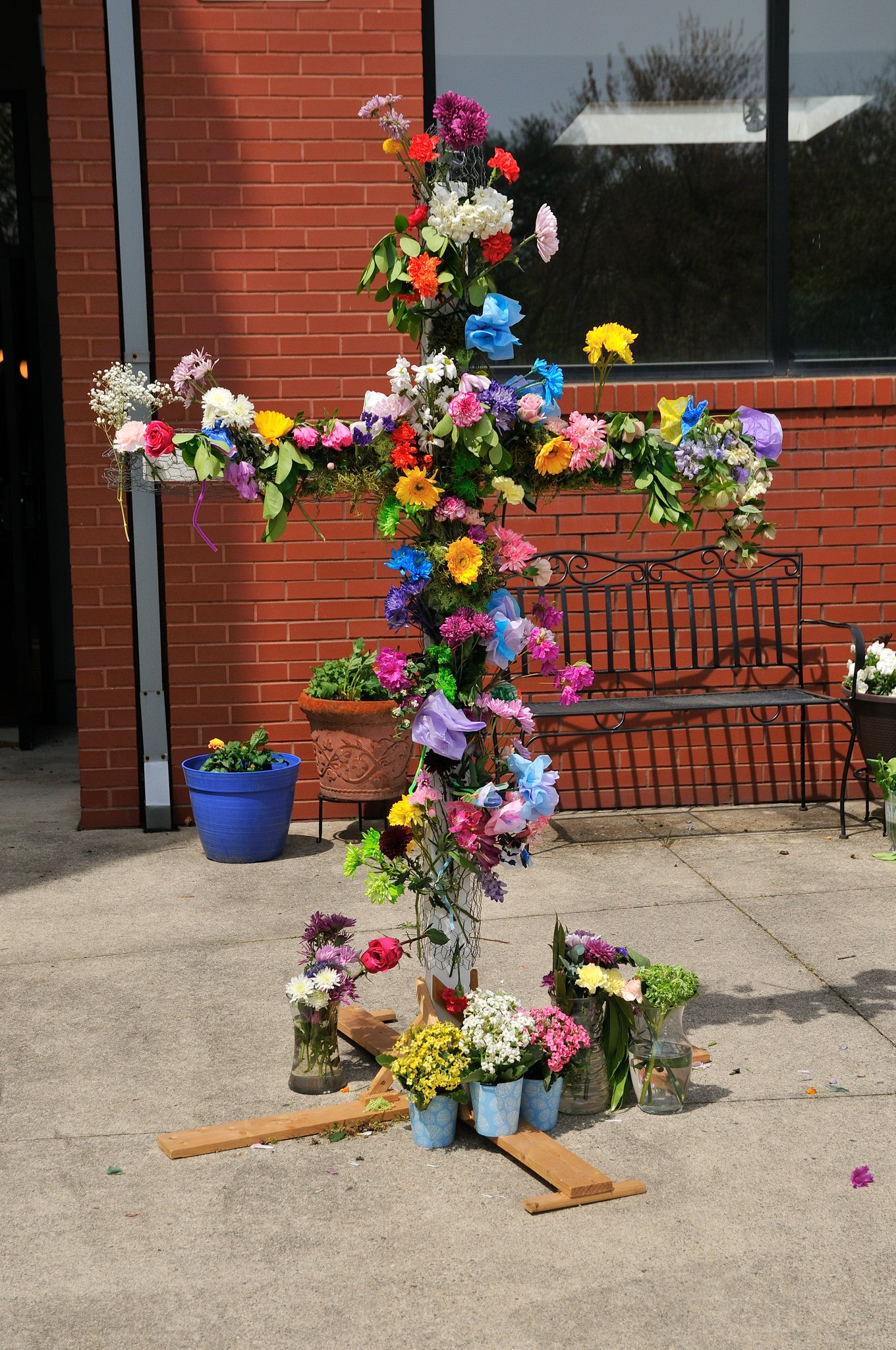 A cross made of flowers stands outdoors on a concrete surface near a brick building with large windows. The cross is decorated with numerous colorful flowers in pots and vases, and there is a wooden base supporting it.