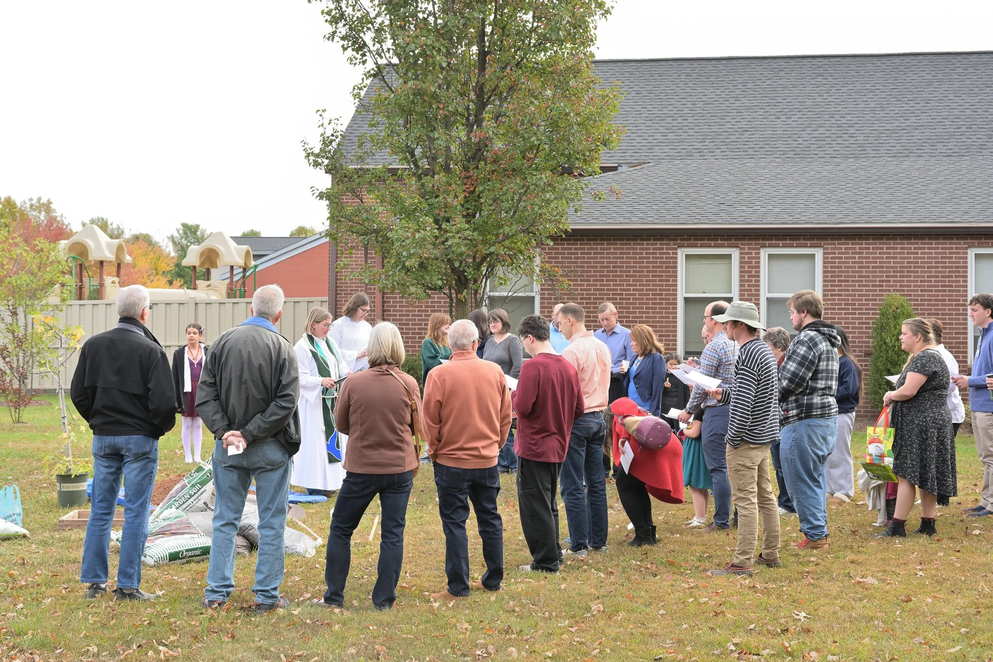 A group of people gathered outdoors on a lawn in front of a brick house with a tree, listening to a speaker. Some are holding papers, and gardening supplies are at the side.