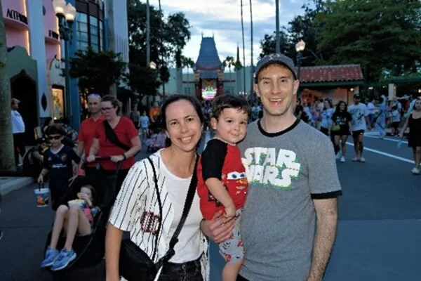 Family of three posing at a theme park, with a castle in the background. The woman is smiling, the man is holding a young boy, and there are other visitors walking around.