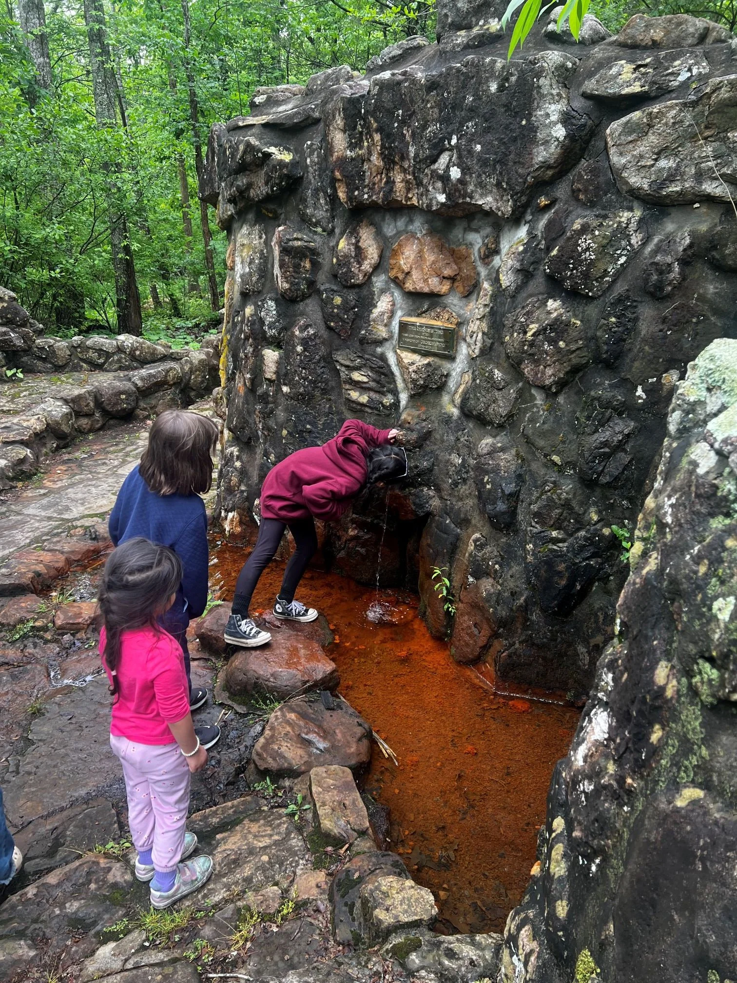 Children exploring a rocky outdoor area with a small stream of water flowing over a stone wall in a forested environment.