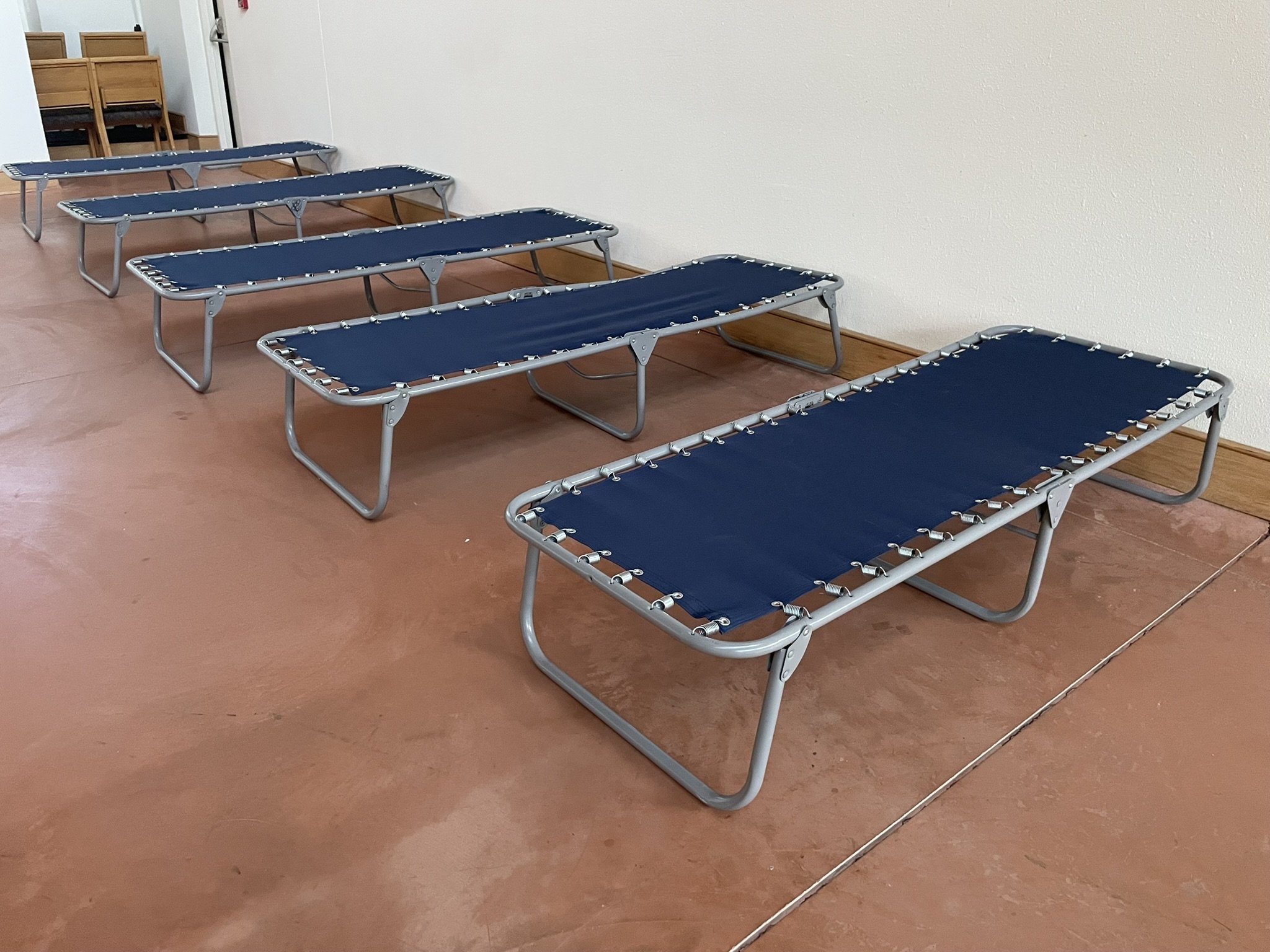 Five blue foldable cots arranged in a row against a white wall in a room with a brown floor and some wooden chairs stacked in the background.