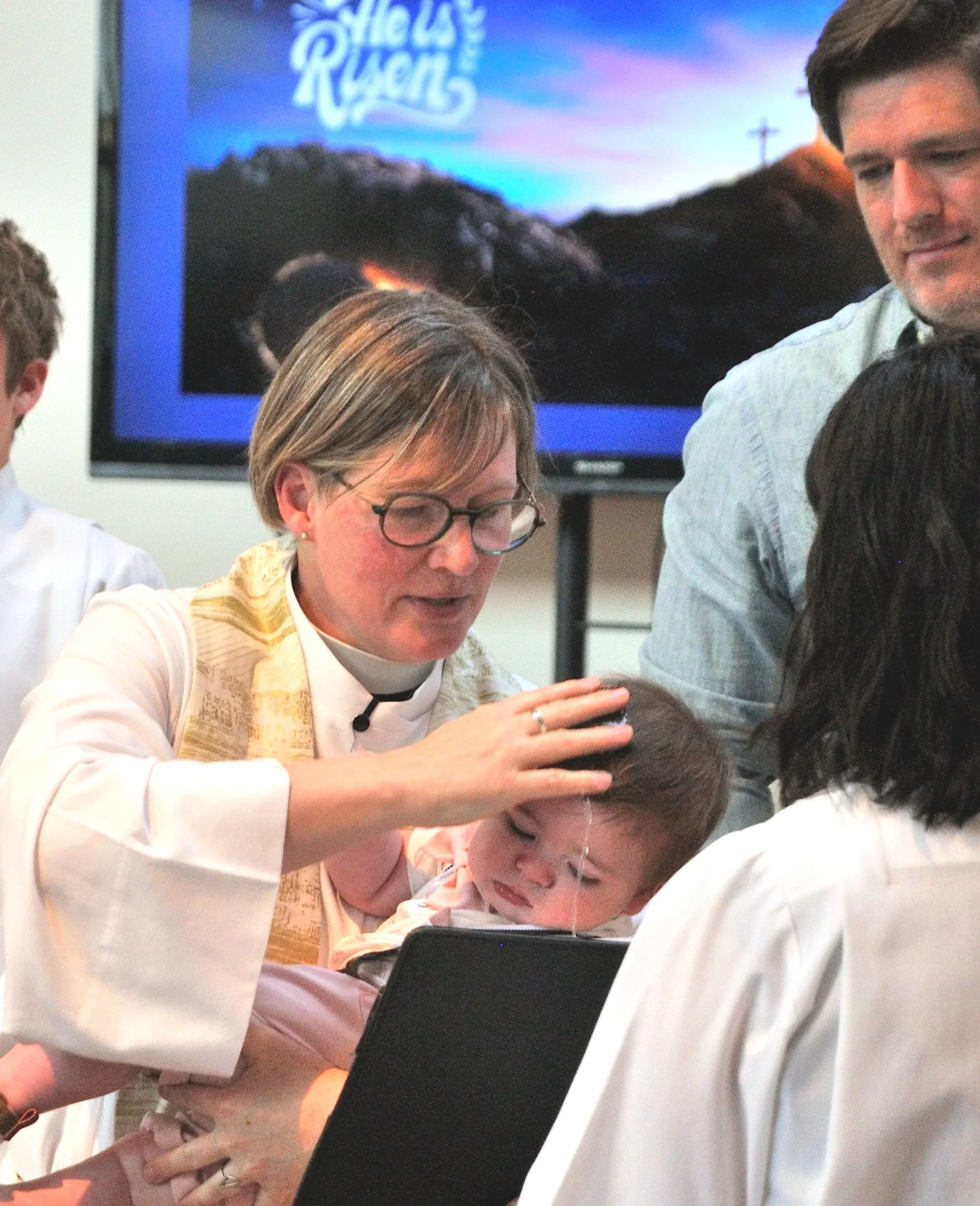 A religious ceremony, with a bishop laying hands on a young person's head during an ordination, in a room with a screen in the background displaying the words 'Vela Racer'.