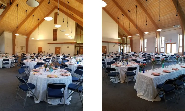 Banquet hall set up for an event with round tables covered in white tablecloths, decorated with flowers, and surrounded by blue chairs under a wooden, vaulted ceiling with hanging lights.