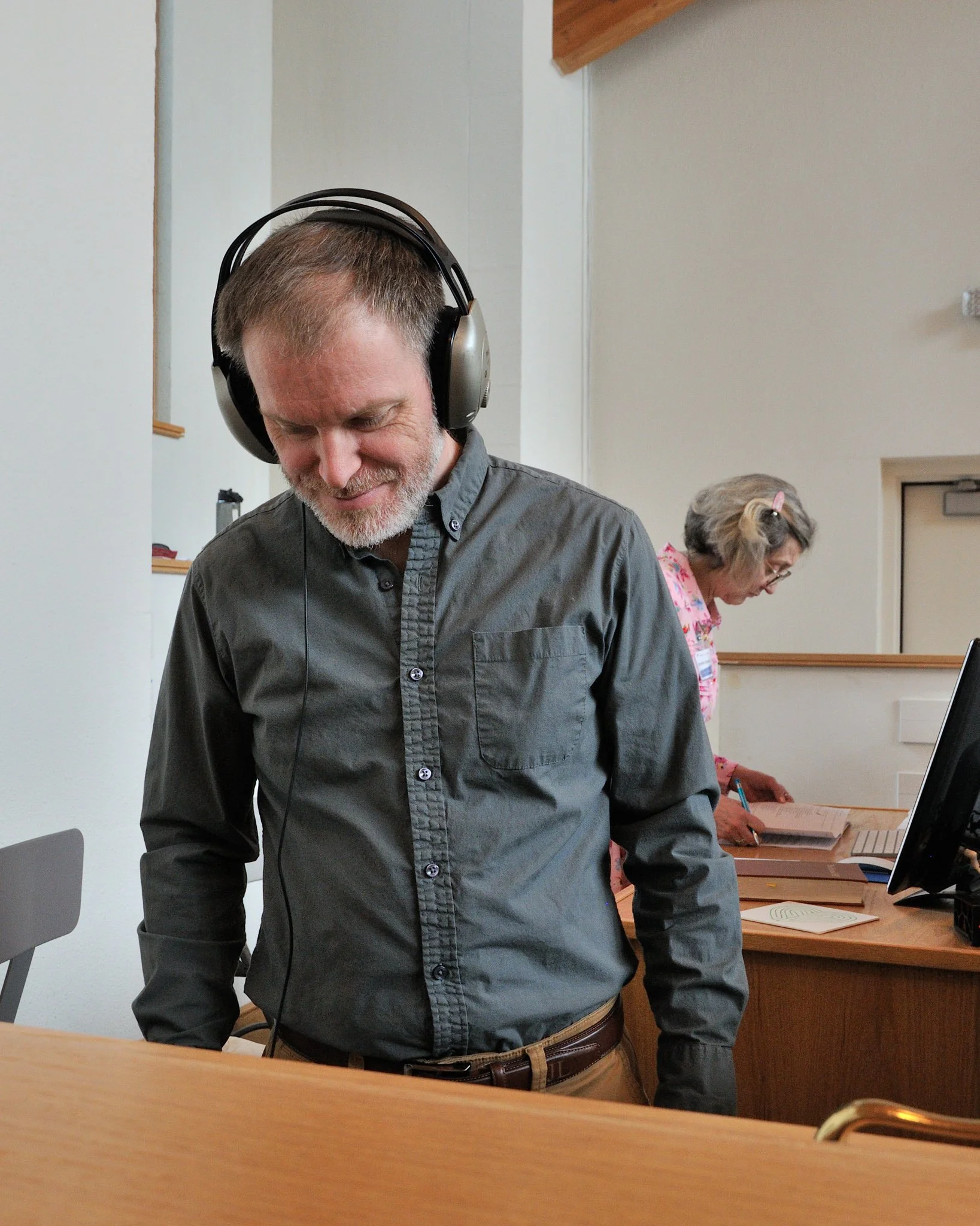 A middle-aged man with gray hair and a beard, smiling and wearing large over-ear headphones, stands in a room with beige walls, a wooden ceiling, and a woman with glasses and blonde hair working at a desk in the background.