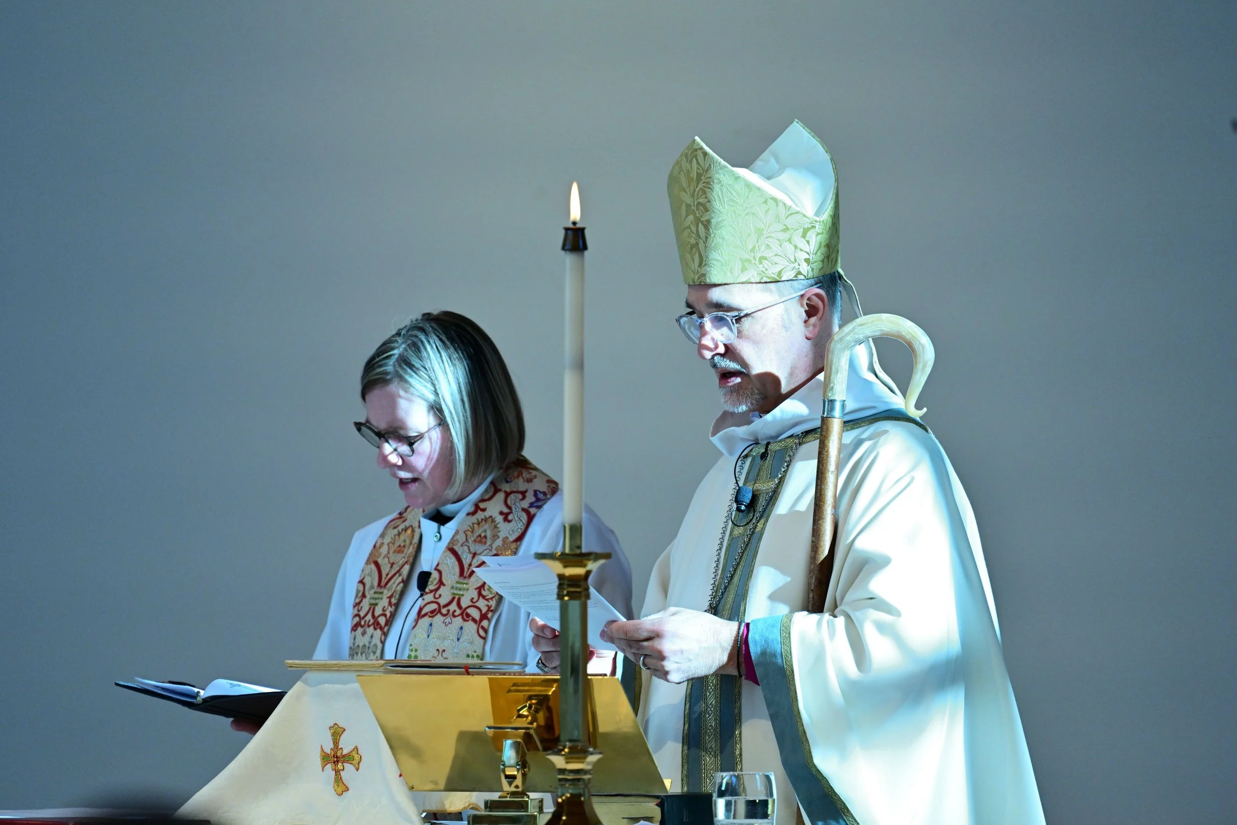 A priest and a woman are standing at an altar during a religious service, reading from papers, with a lit candle and a chalice on the altar.