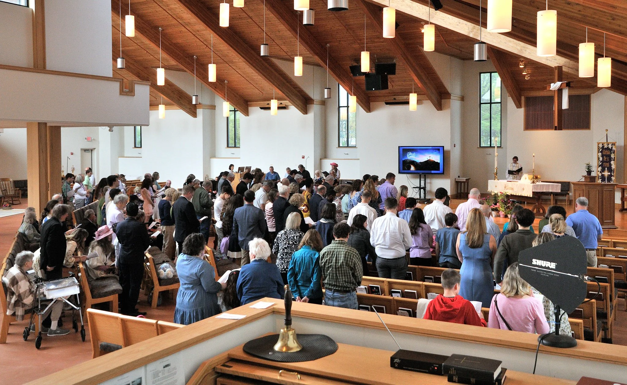 Inside a church during a service with a large congregation of people standing and sitting, facing the altar. The church has wooden accents on the ceiling with hanging lights, large windows, and various religious decorations.