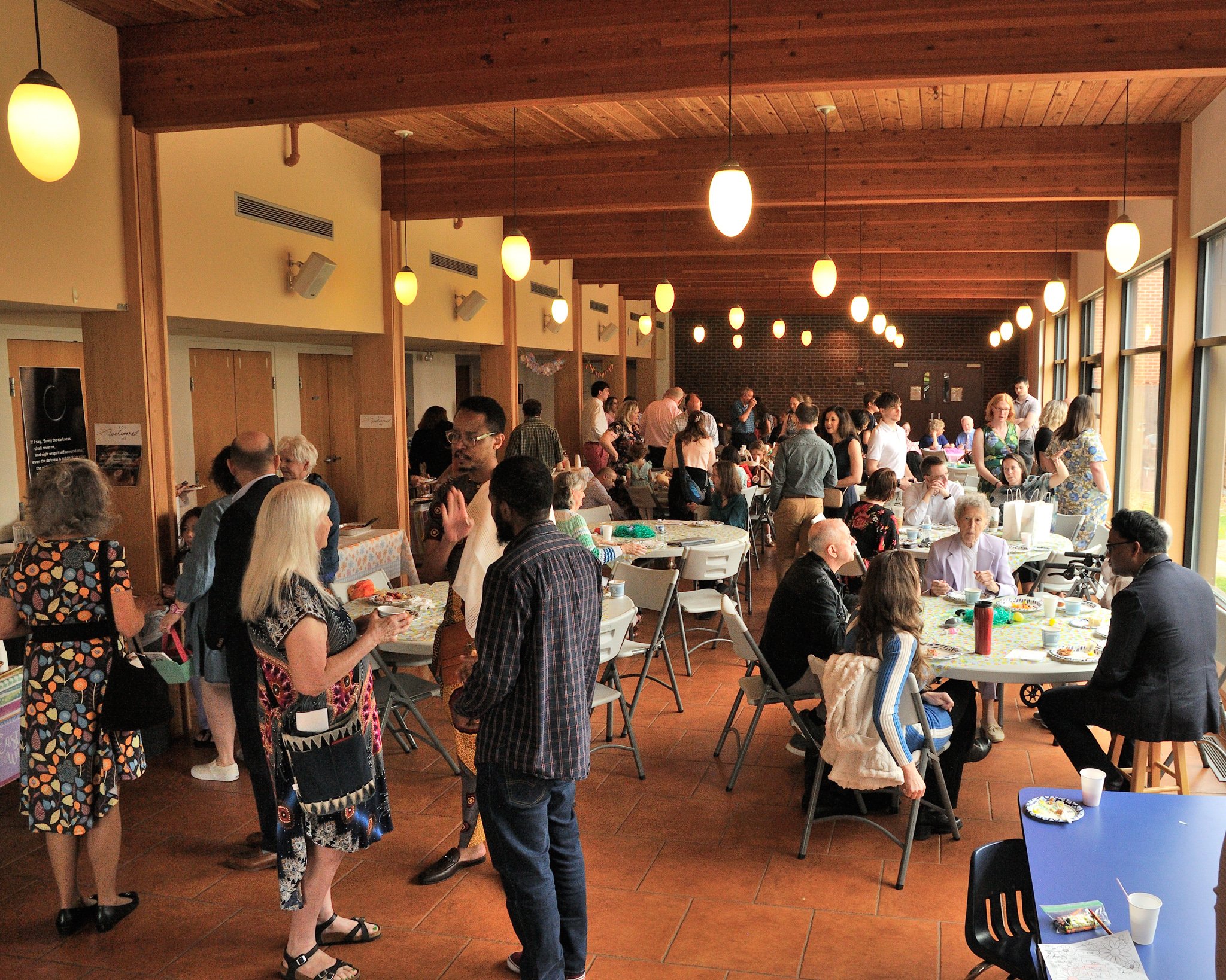 People gathered indoors in a decorated hall with wooden ceiling panels and large windows, socializing and engaging in activities around tables.