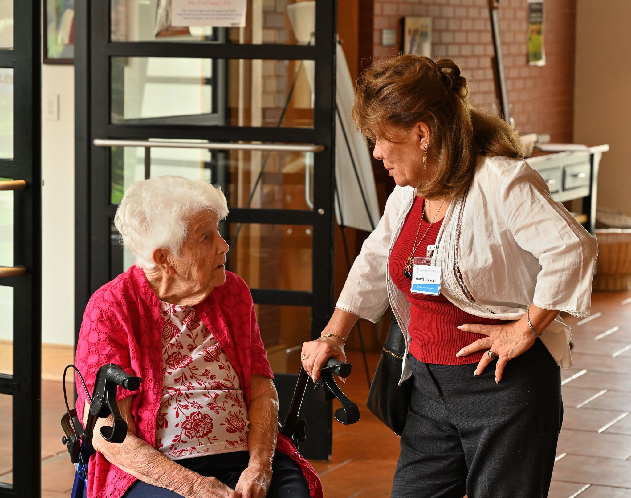 Gloria Jackson is talking to an elderly woman with white hair, who is seated in a wheelchair. The elderly woman is wearing a red jacket over a patterned blouse. They are in an indoor setting with brick walls, large windows, and some artwork or poster