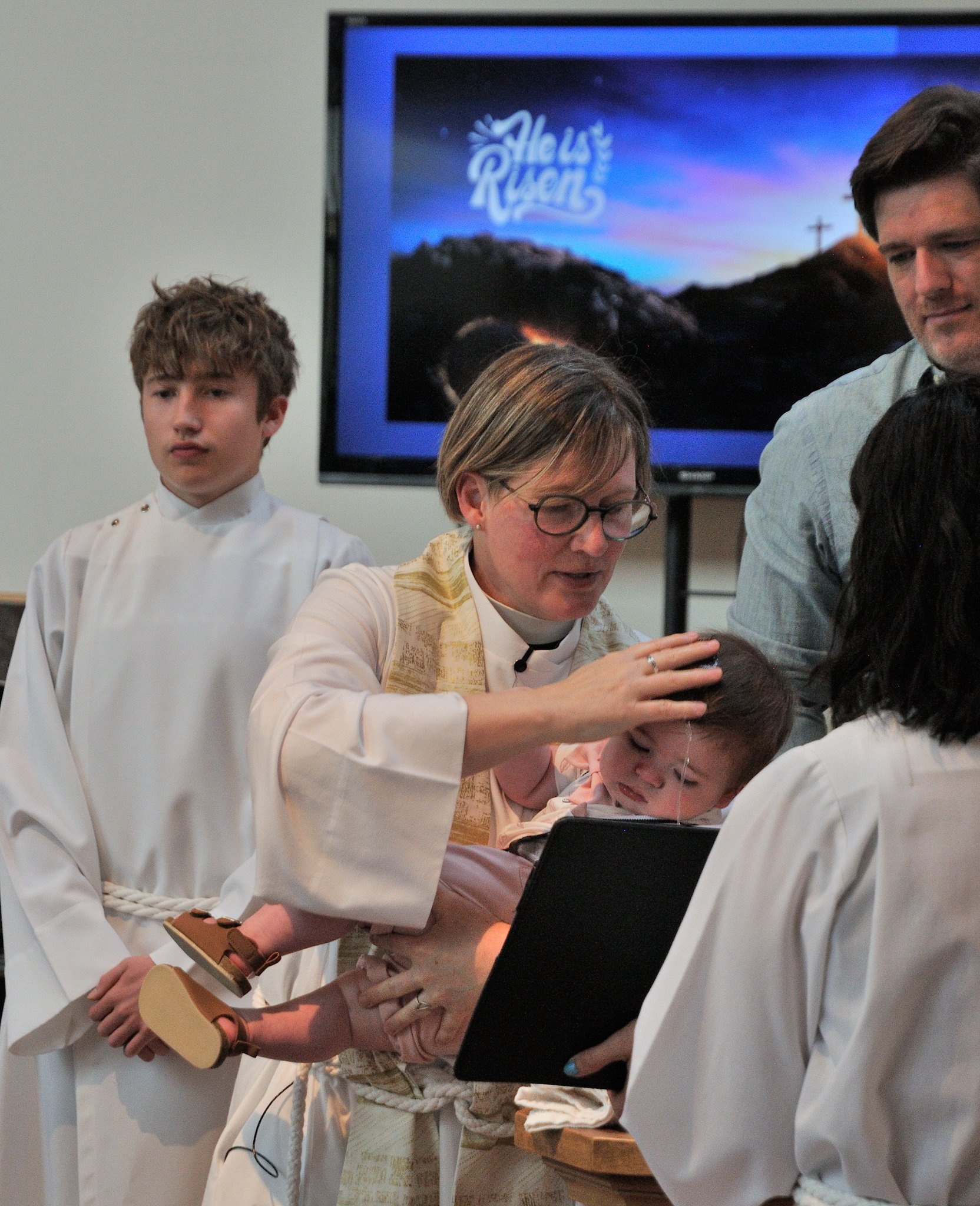 Group of people dressed in white robes, with one person holding a small guitar, participating in a religious ceremony or rehearsal with a woman performing a ritual or prayer, in a room with a monitor displaying a logo for 'He Is Risen' in the backgro