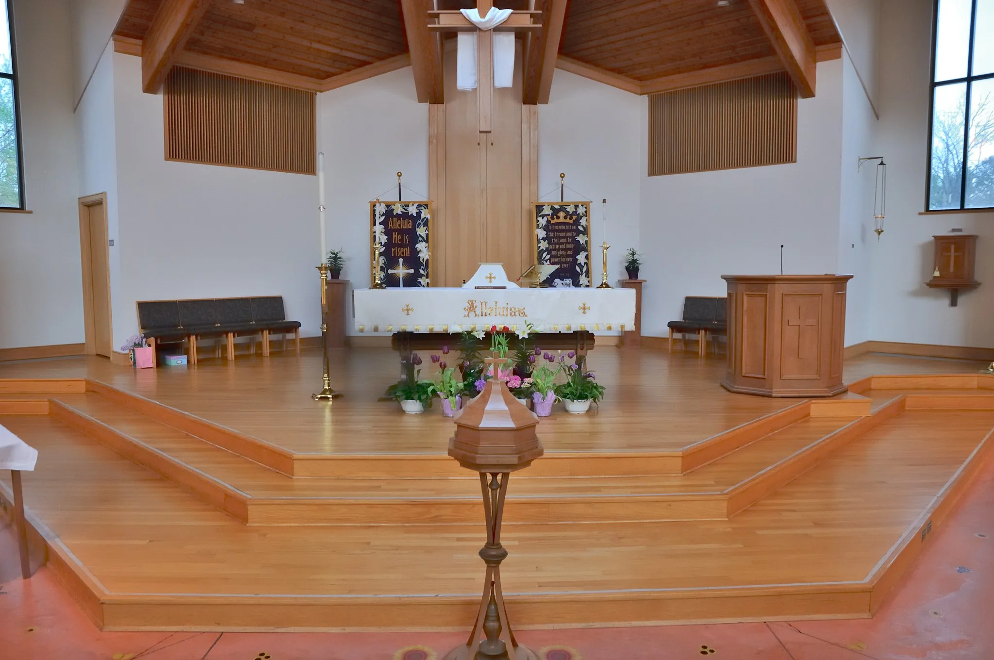 Interior view of a church sanctuary with an altar draped in white cloth, floral arrangements, and banners that read 'Alleluia He is Risen' and a prayer of praise. There are black chairs along the sides, wooden steps leading to the altar, and large wi