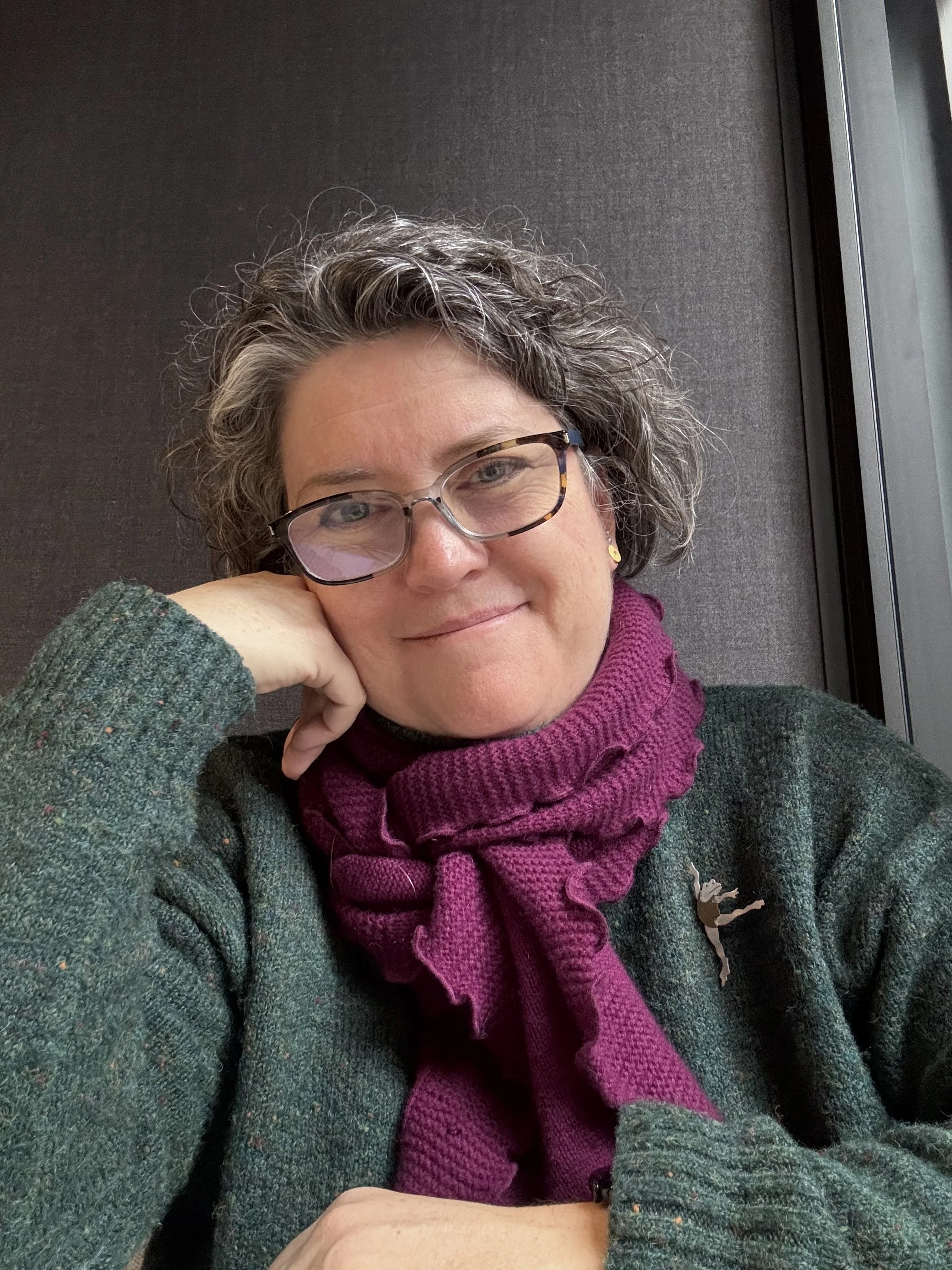 A woman with curly gray hair wearing glasses, a green sweater, and a purple scarf, resting her head on her hand and smiling, sitting by a window with a dark background.