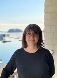 Woman with shoulder-length dark hair and glasses standing outside near water, smiling.