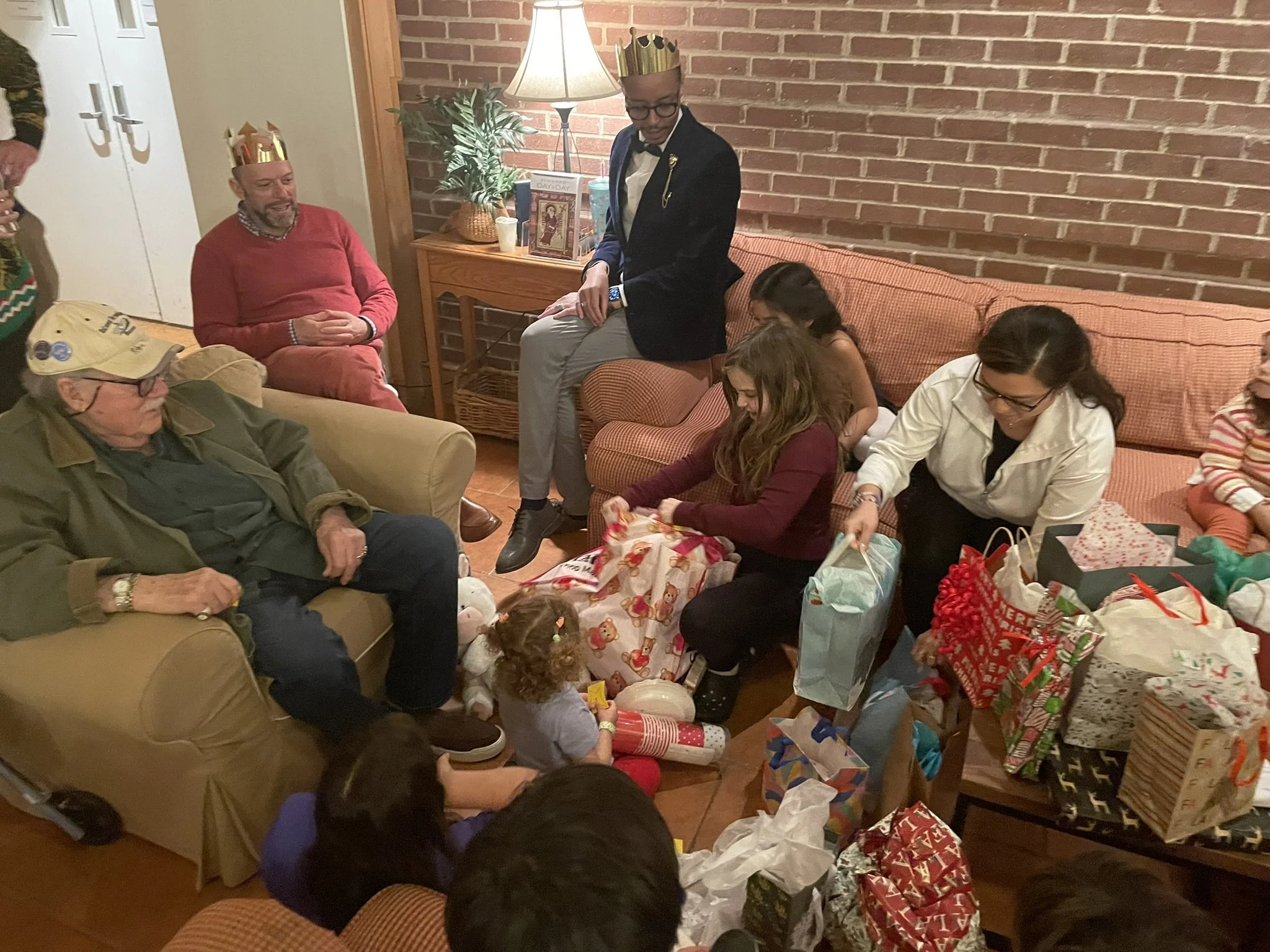 People sitting on couches and chairs in a living room, opening Christmas presents with party hats, gifts, and wrapping paper scattered around, a brick wall background, and a lamp on a side table.