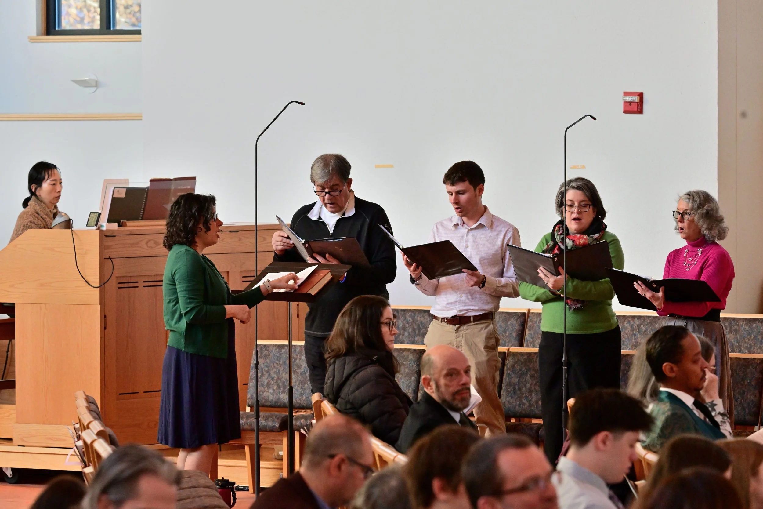 People standing and singing during a choir rehearsal or performance in a church or auditorium. The group includes men and women holding sheet music, with a woman in a green sweater standing at a music stand, and a man in a black jacket with glasses r