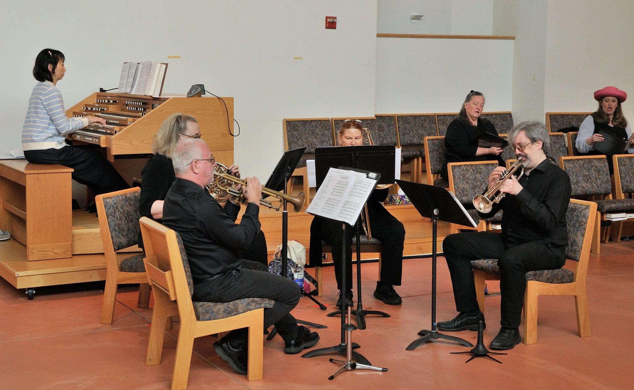 A group of musicians practicing in a church sanctuary, including a young girl on an organ, and five adults playing brass and wind instruments, with music stands in front of them.