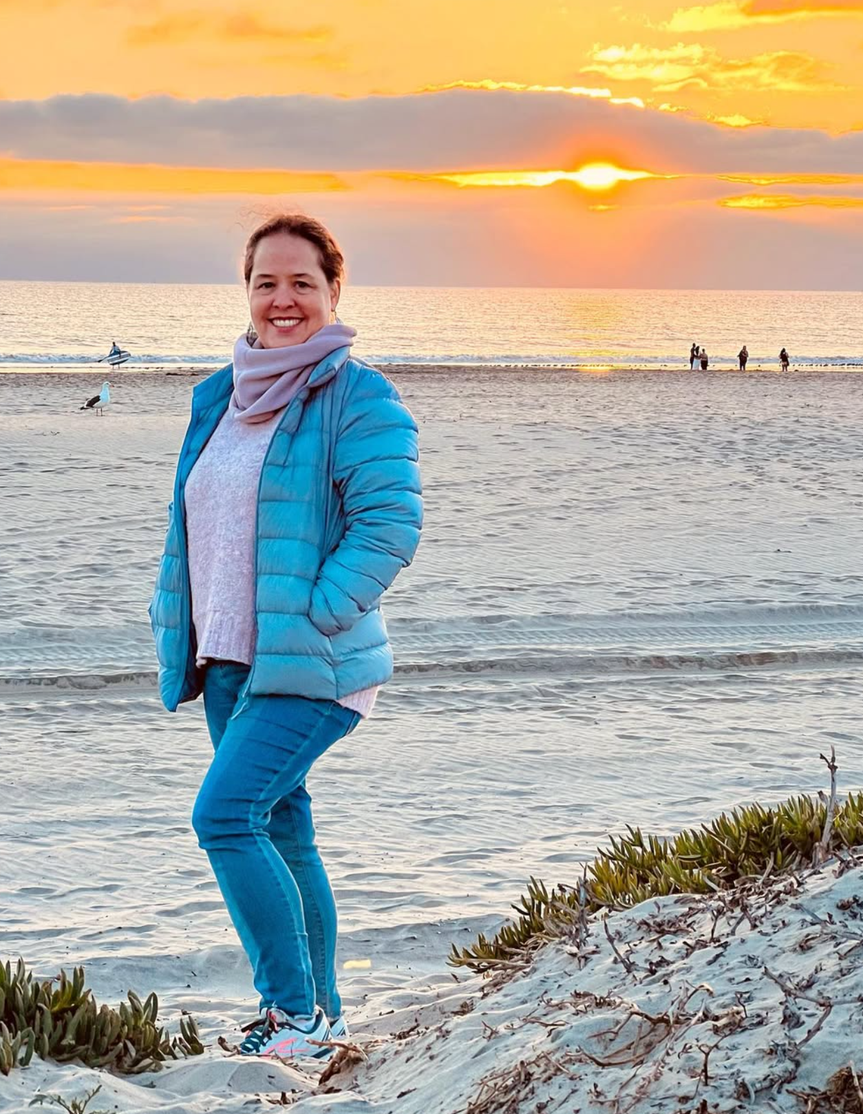 A woman smiling on a beach during sunset, wearing a blue puffer jacket, light pink scarf, and blue pants, with greenery in the foreground and the ocean and other beachgoers in the background.